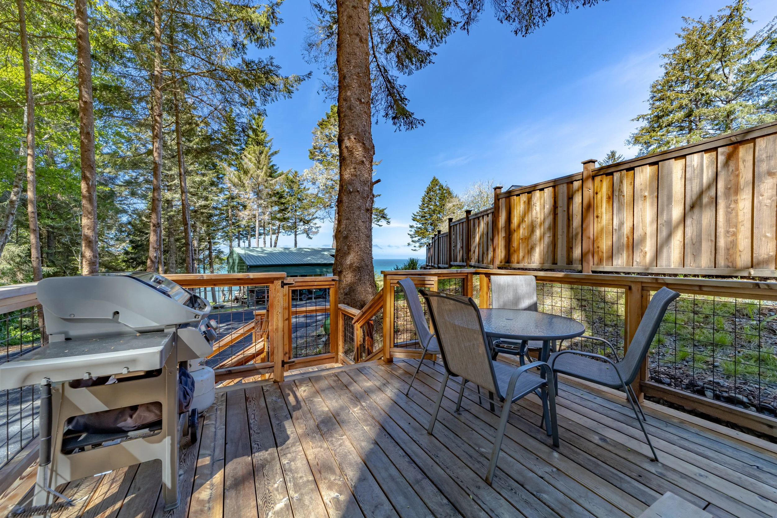 Wooden deck with outdoor furniture including a round table and four chairs, a gas grill, surrounded by trees and a wooden privacy fence with a view of the sky and distant trees.