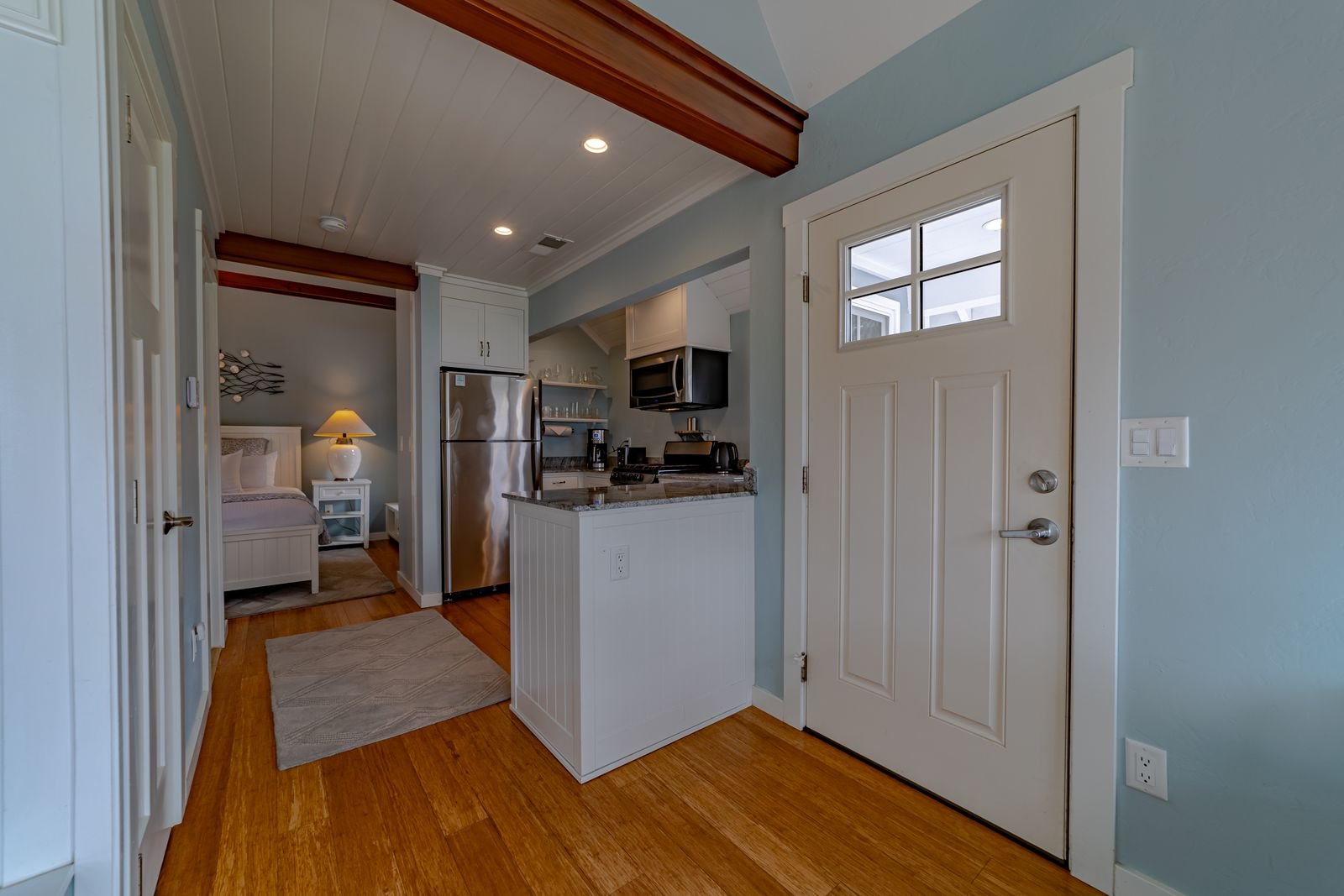 Interior of a small apartment with a kitchenette, a doorway, and a bedroom visible in the background, featuring hardwood floors, light blue walls, and white trim.