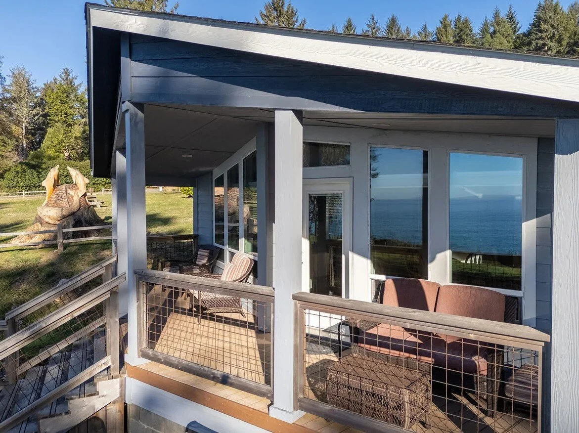 A balcony with outdoor furniture, including a brown sofa, wicker chairs, and an outdoor table, overlooking a body of water with trees in the background.