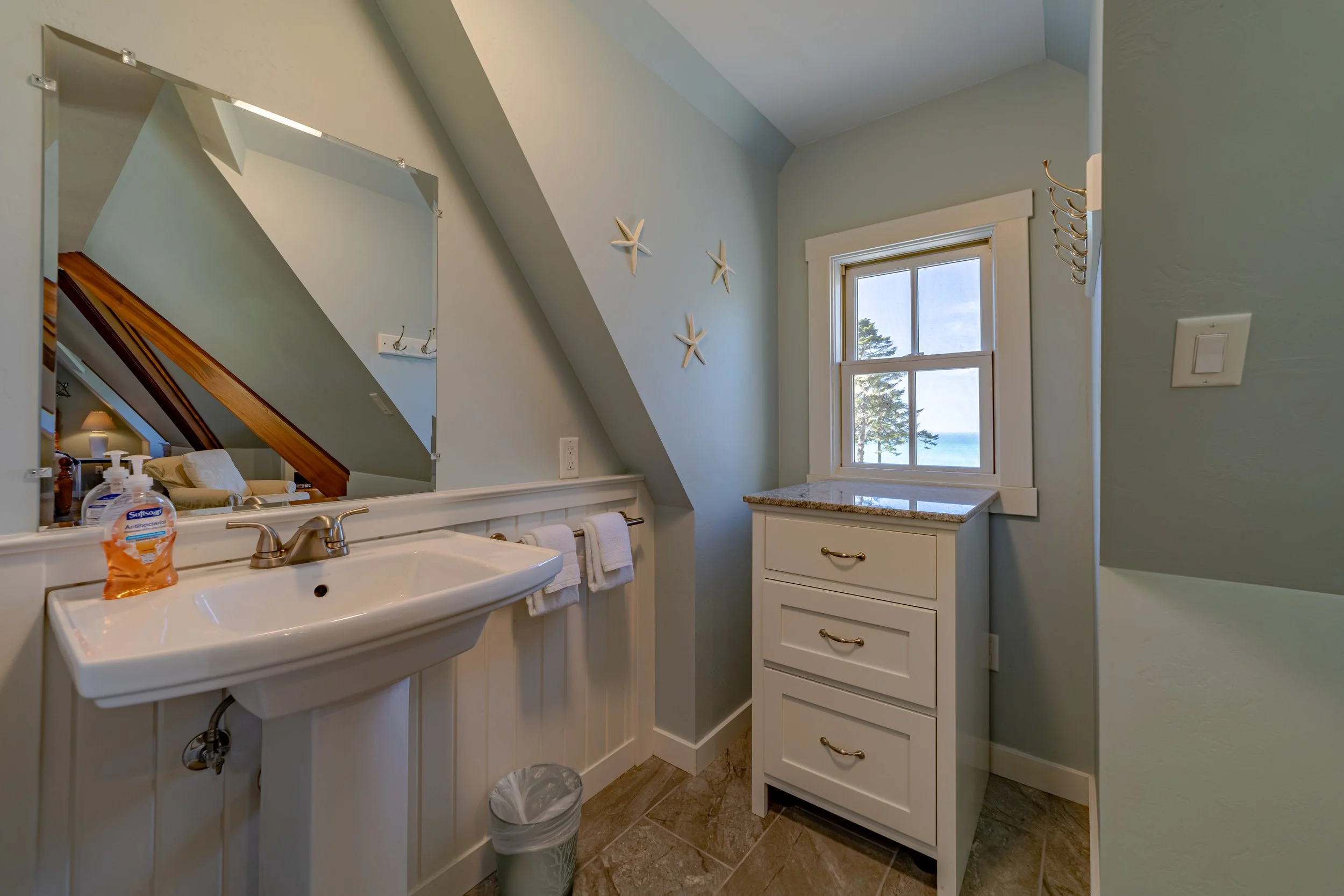 Bathroom with a white sink, a mirror, a small white cabinet, a window showing trees and the ocean, decorative starfish on the wall, and a trash can on the floor.