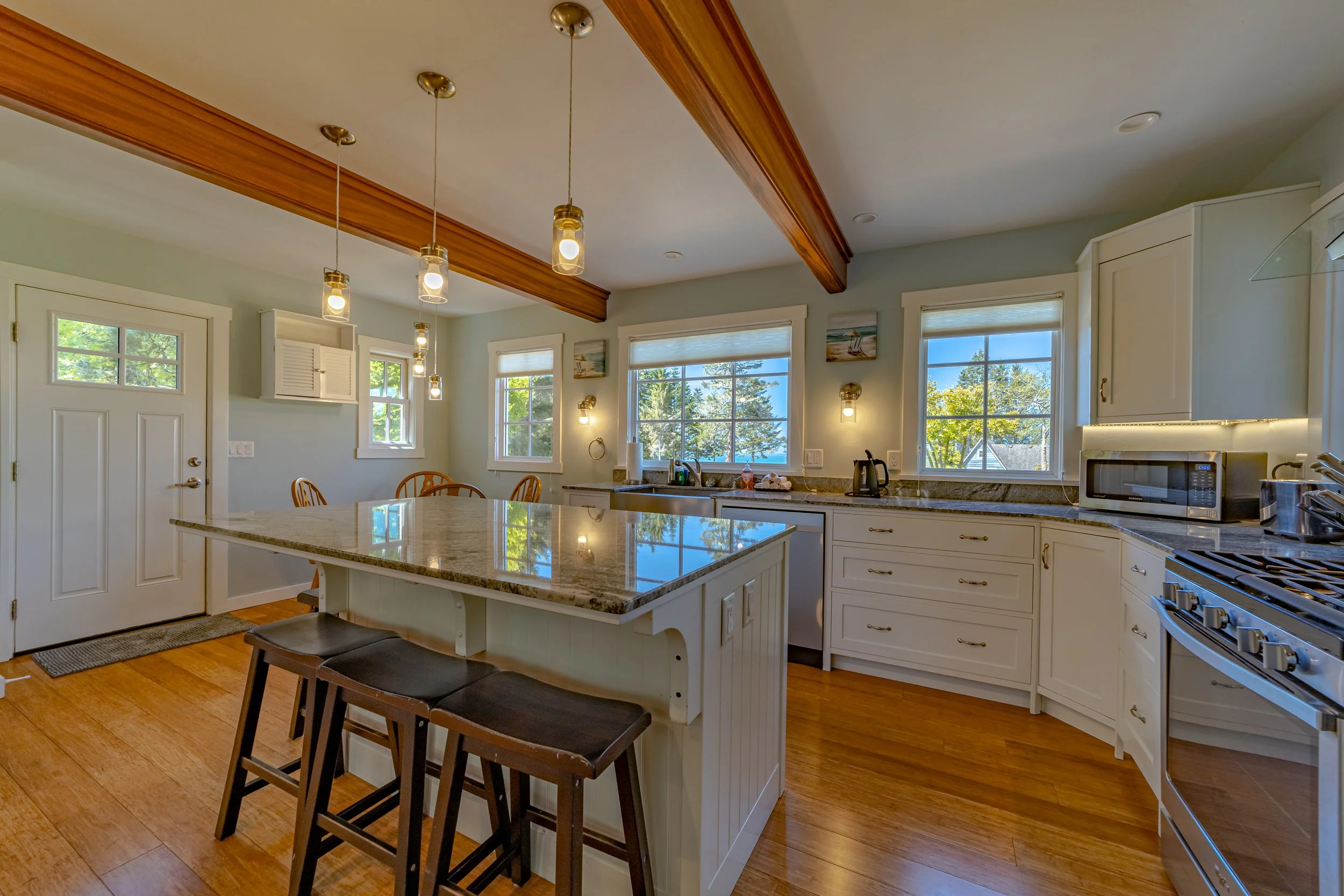 Bright kitchen with white cabinets, granite countertops, and large windows showing outdoor trees; island with seating, pendant lights hanging from wooden beam ceiling.
