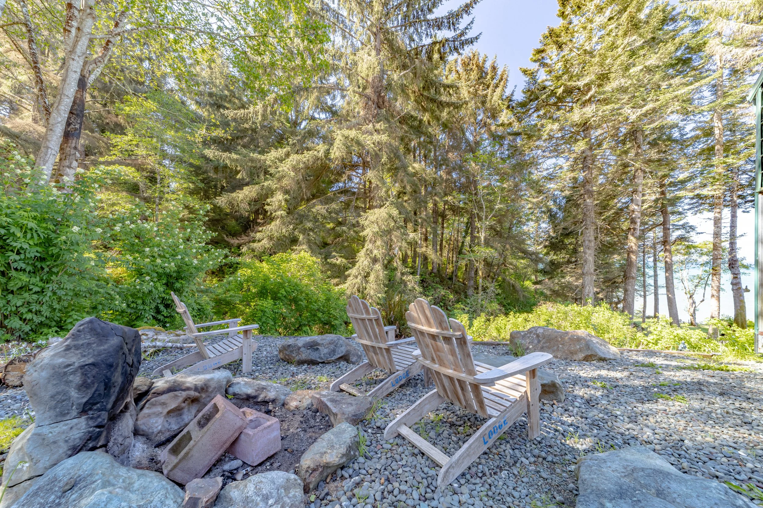 Three wooden Adirondack chairs on a rocky outdoor area surrounded by large rocks and green trees, with a clear blue sky above.