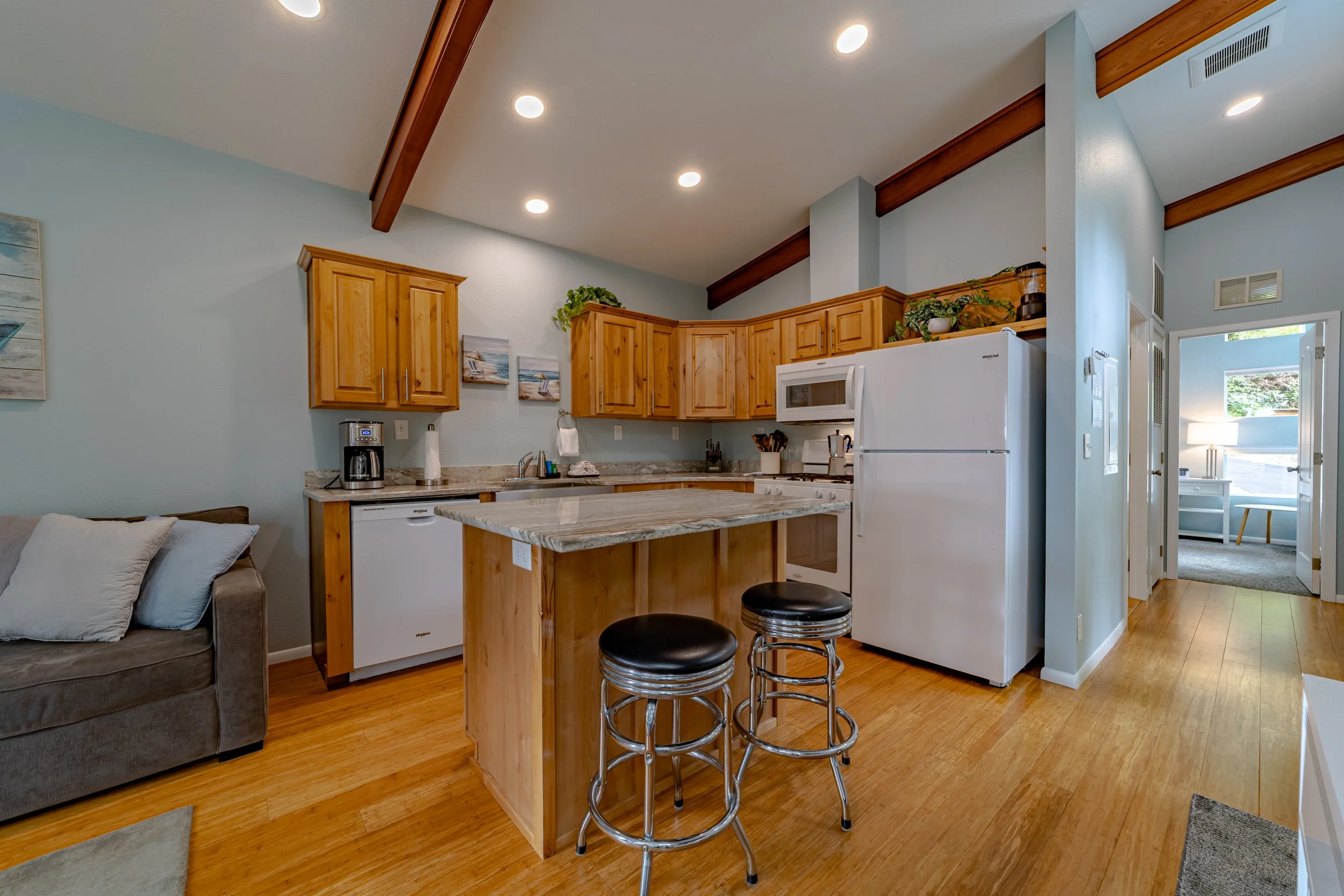 Kitchen with wooden cabinets, white appliances, granite countertops, a kitchen island with two black barstools, and a connecting living space with a gray sofa.