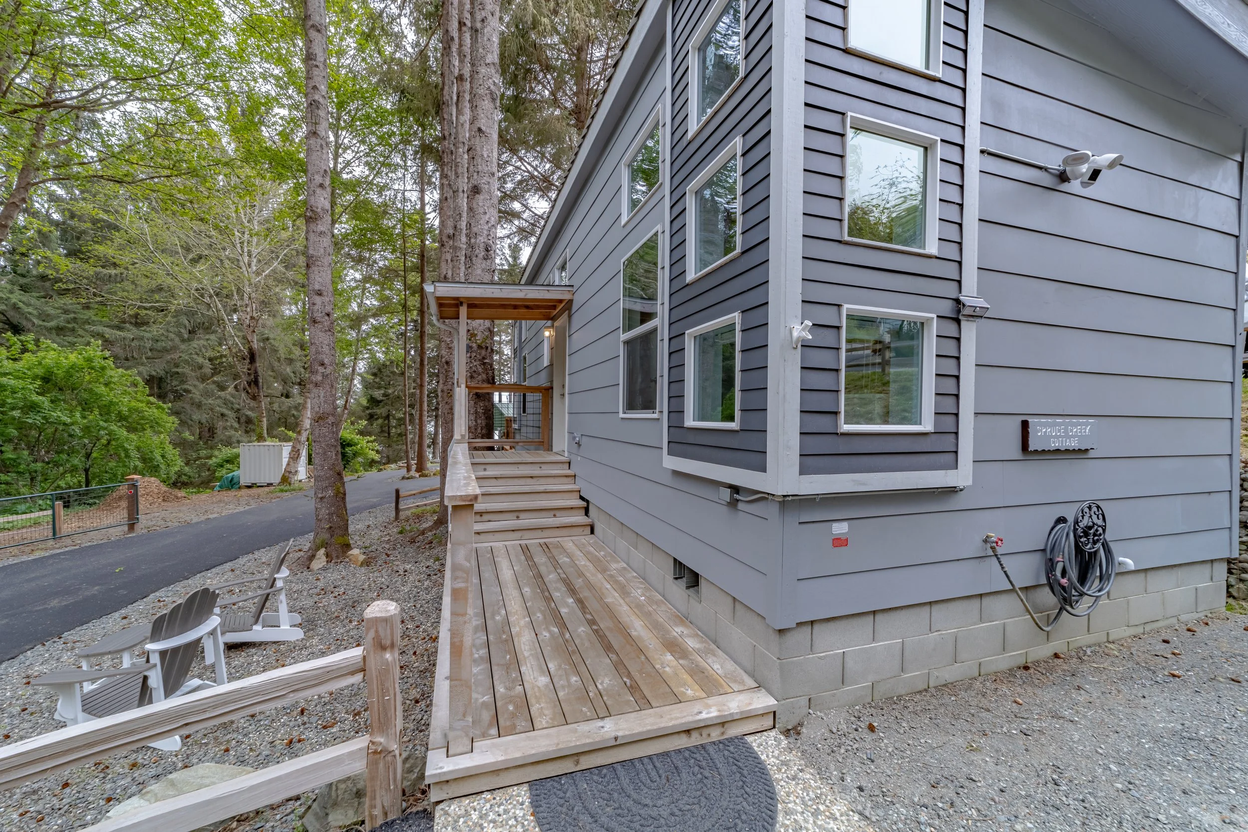 Side view of a modern gray house with multiple windows, a small wooden deck, and an outdoor hose reel on the wall. Surrounding tall trees and a paved pathway with outdoor chairs.