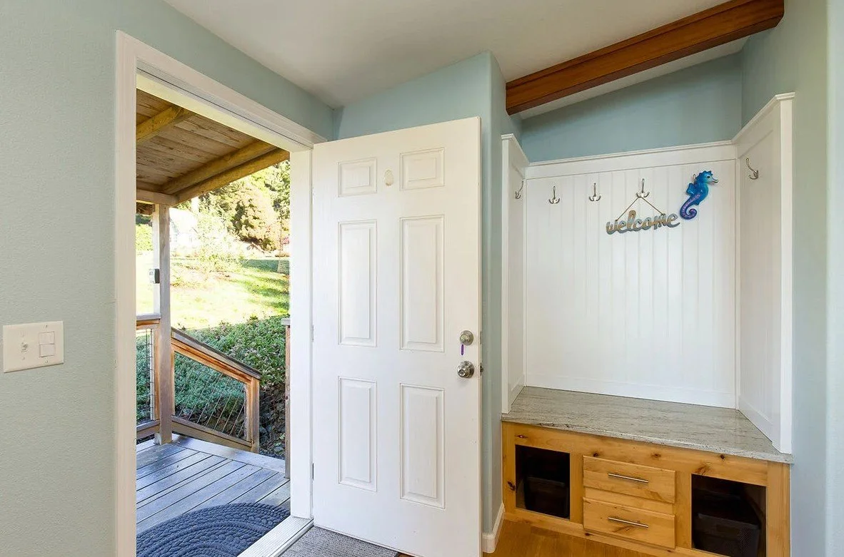 Interior entryway with open front door leading outside porch, light blue walls, white wainscoting with hooks, decorative "welcome" sign, wooden button frame, and a small cabinet with drawers and open shelves.