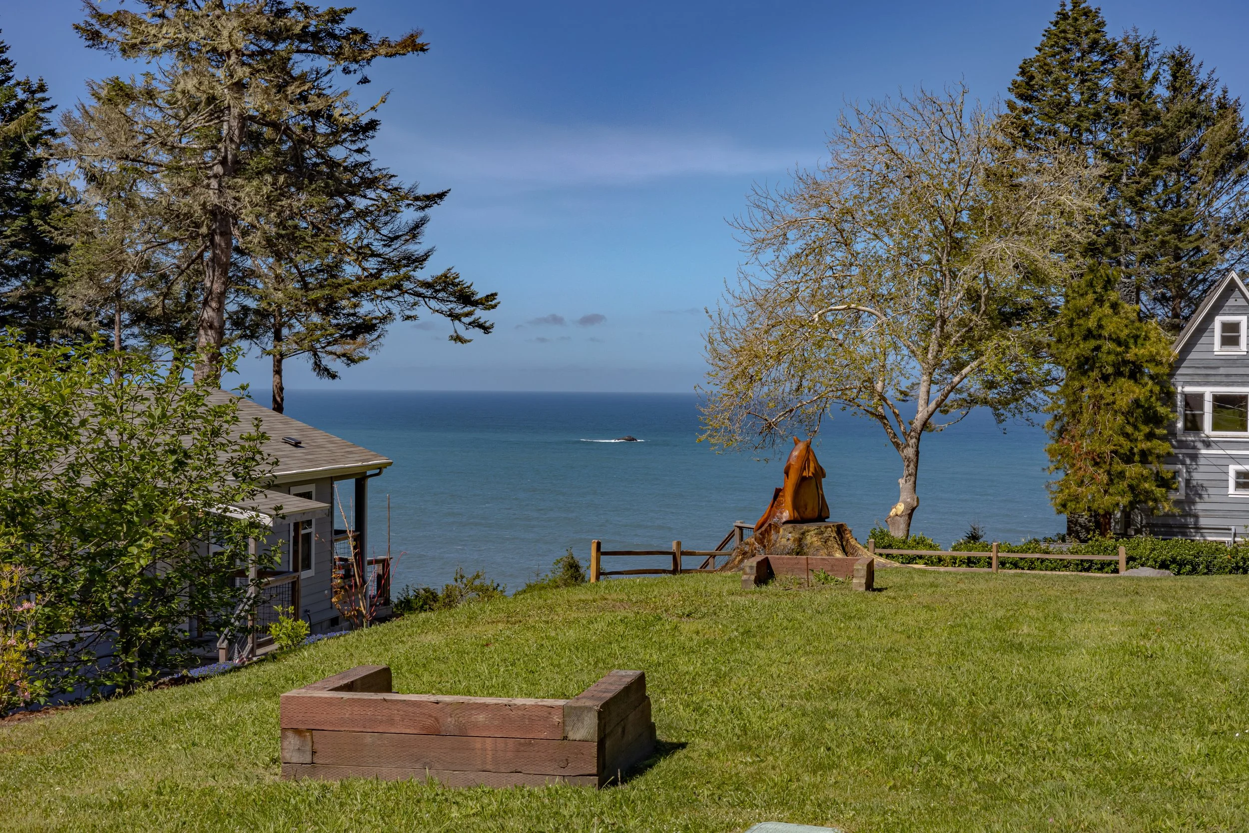 A scenic view of a grassy yard overlooking the ocean with trees and houses on either side, and a sculpture of a person sitting on a tree stump in the center.