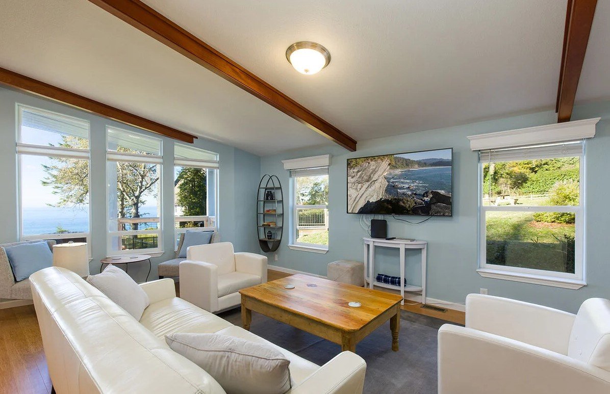 Living room with large windows showing outdoor trees and ocean view, white sofa and armchairs, wooden coffee table, wall-mounted flat-screen TV, smaller table with books, and light blue walls.