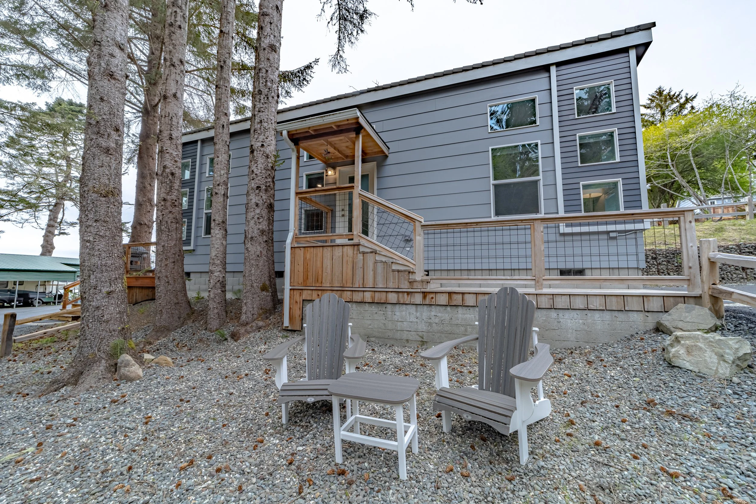 Gray house with a wooden deck and outdoor seating area on a gravel ground, surrounded by tall trees.