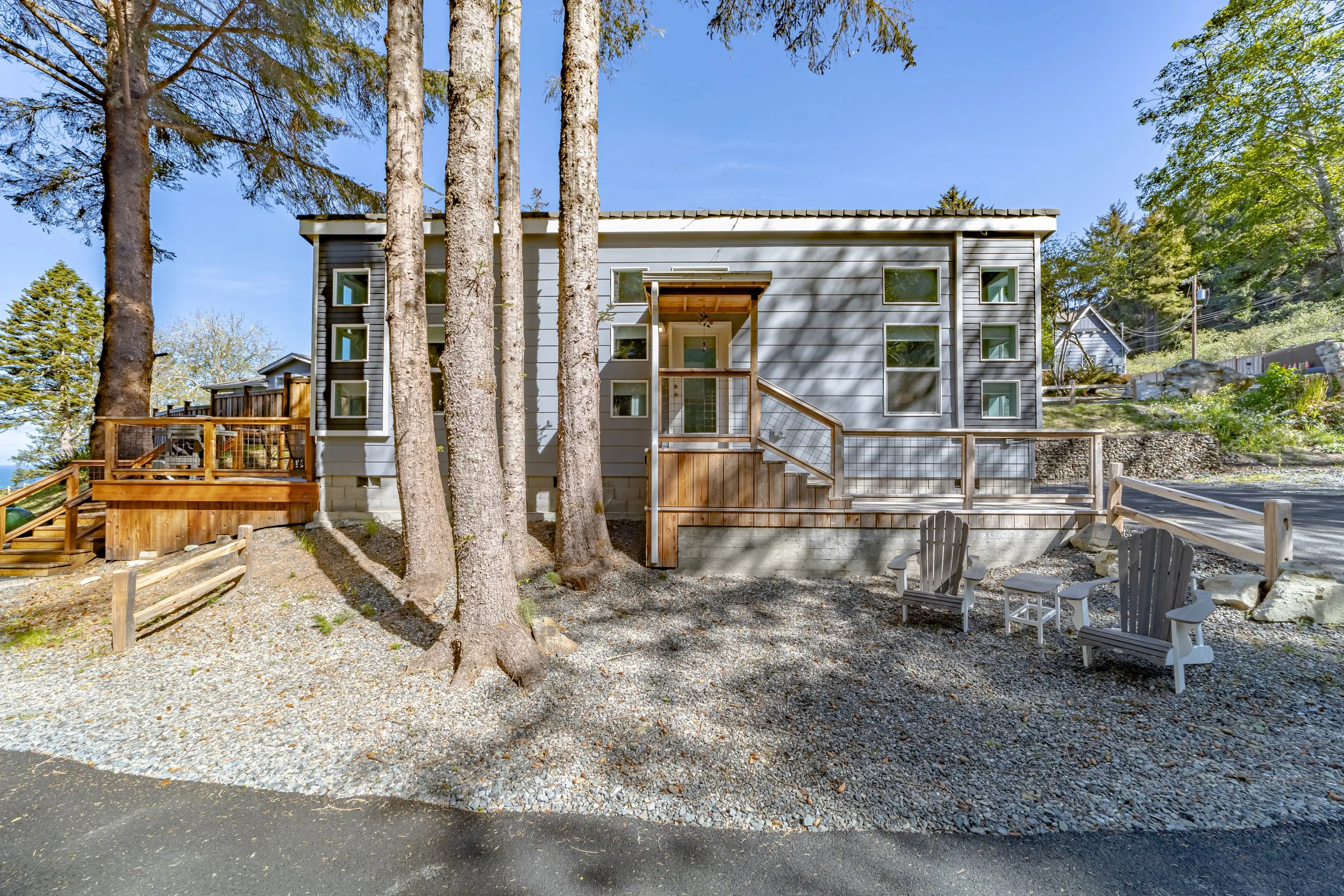 A modern, multi-story house with gray siding, multiple small windows, and a wooden deck in a forested area. There are trees in front, gravel landscaping, and outdoor furniture.