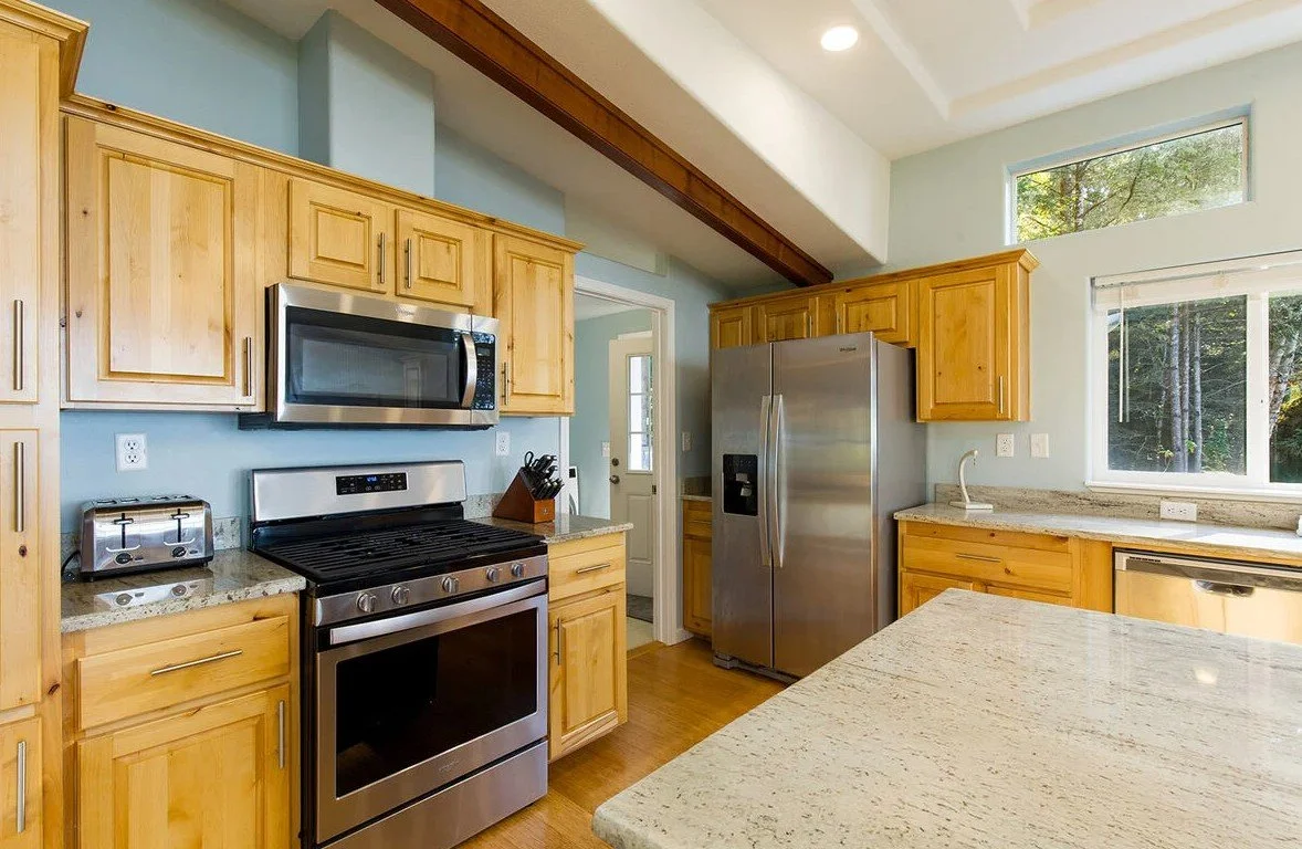 Kitchen with wooden cabinets, stainless steel appliances including a refrigerator, oven, microwave, and dishwasher, granite countertops, and a window with a view of trees outside.