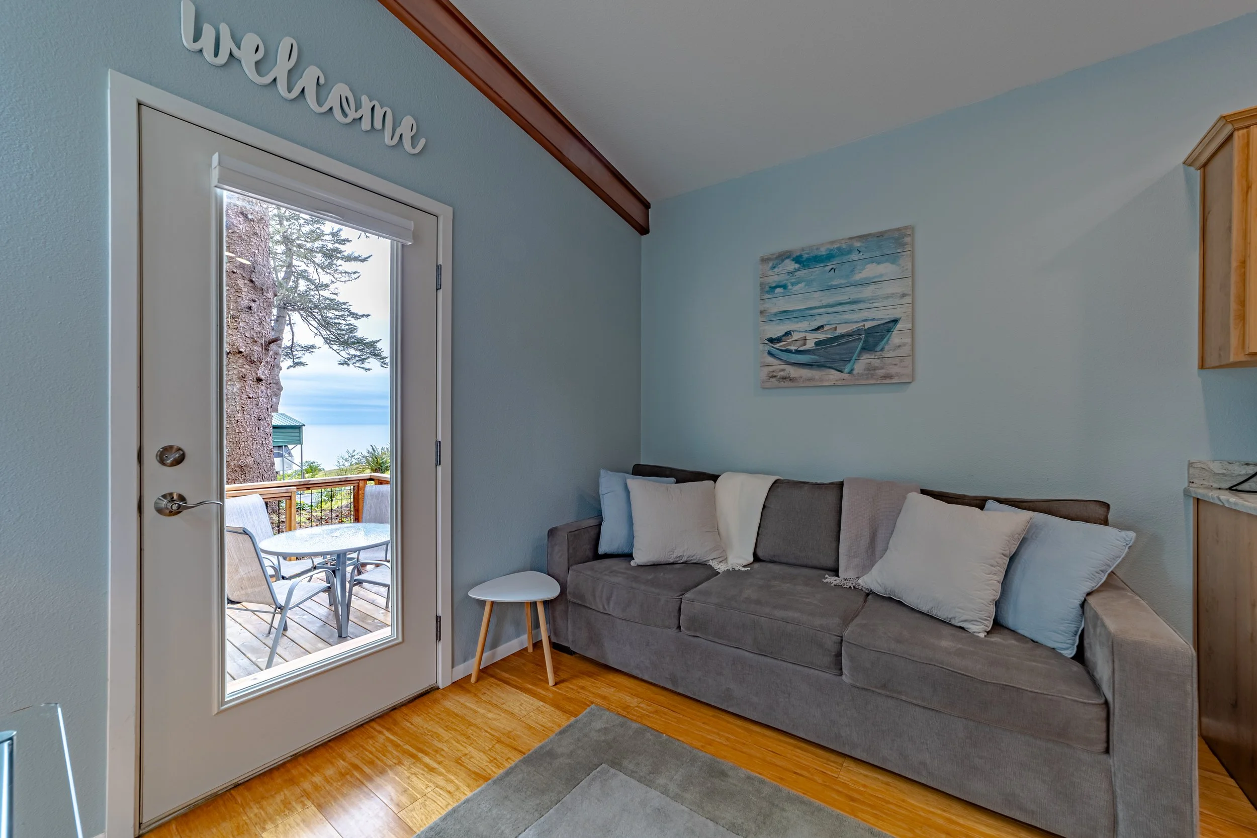Living room with a gray couch topped with multiple pillows, a small white side table, wall artwork of a boat, and a glass door leading to a porch with outdoor seating and a view of trees and water.
