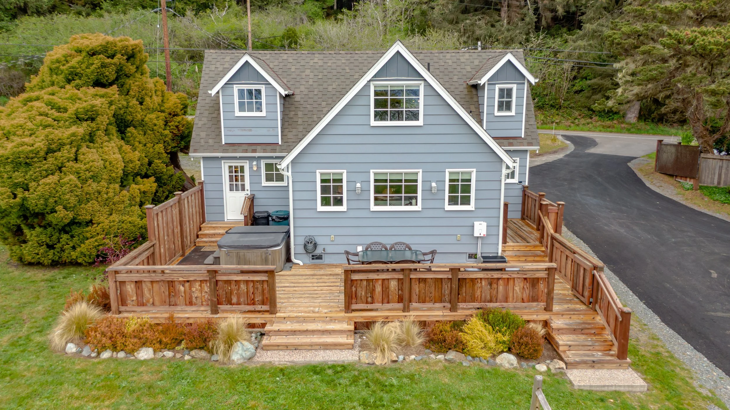 A two-story blue house with a wooden deck in the backyard, featuring outdoor furniture, a hot tub, and a paved driveway, surrounded by trees and greenery.