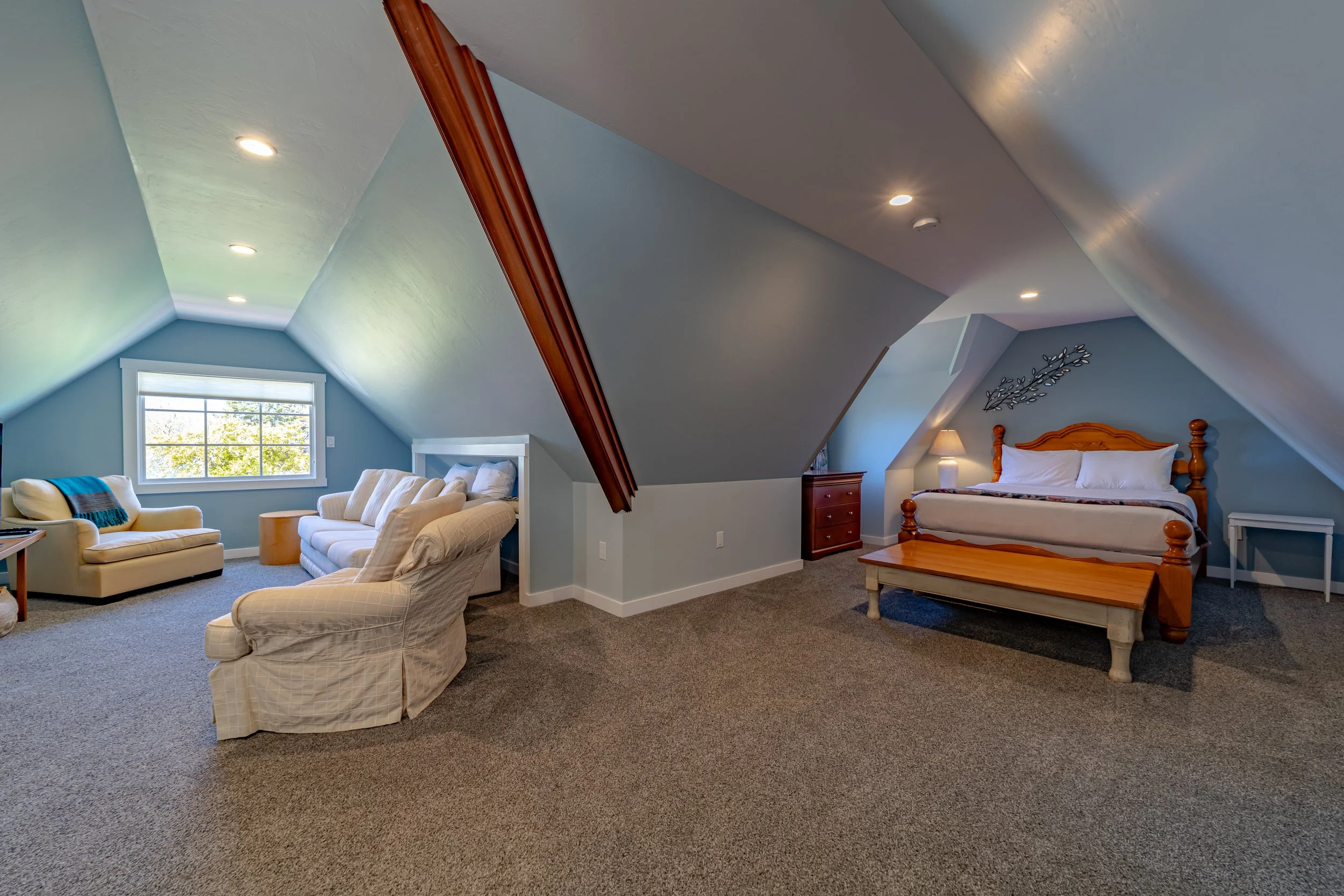 Loft bedroom with sloped ceilings, blue walls, and carpeting. Features a sitting area with beige and white furniture, a large window, and a separate sleeping area with a wooden bed, nightstand, and lamp.