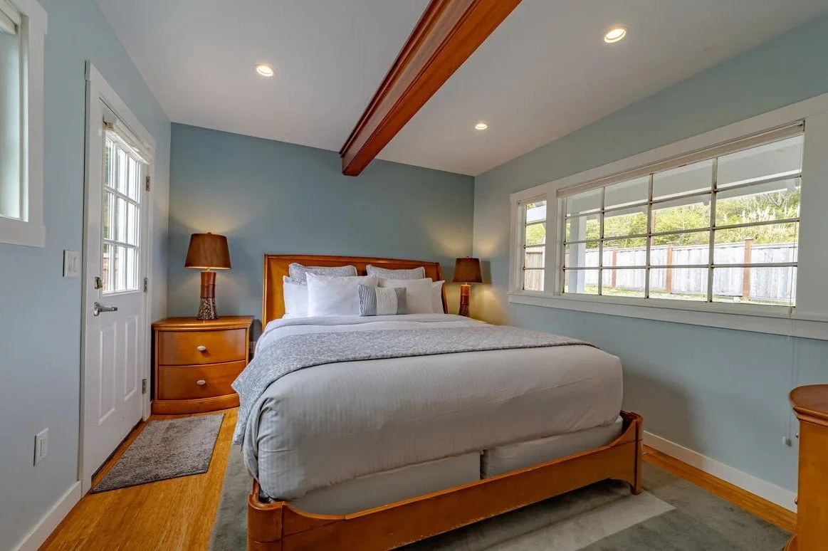A bedroom with a wooden bed frame, white bedding, two bedside tables with lamps, a large window with multiple panes, and a light blue wall.