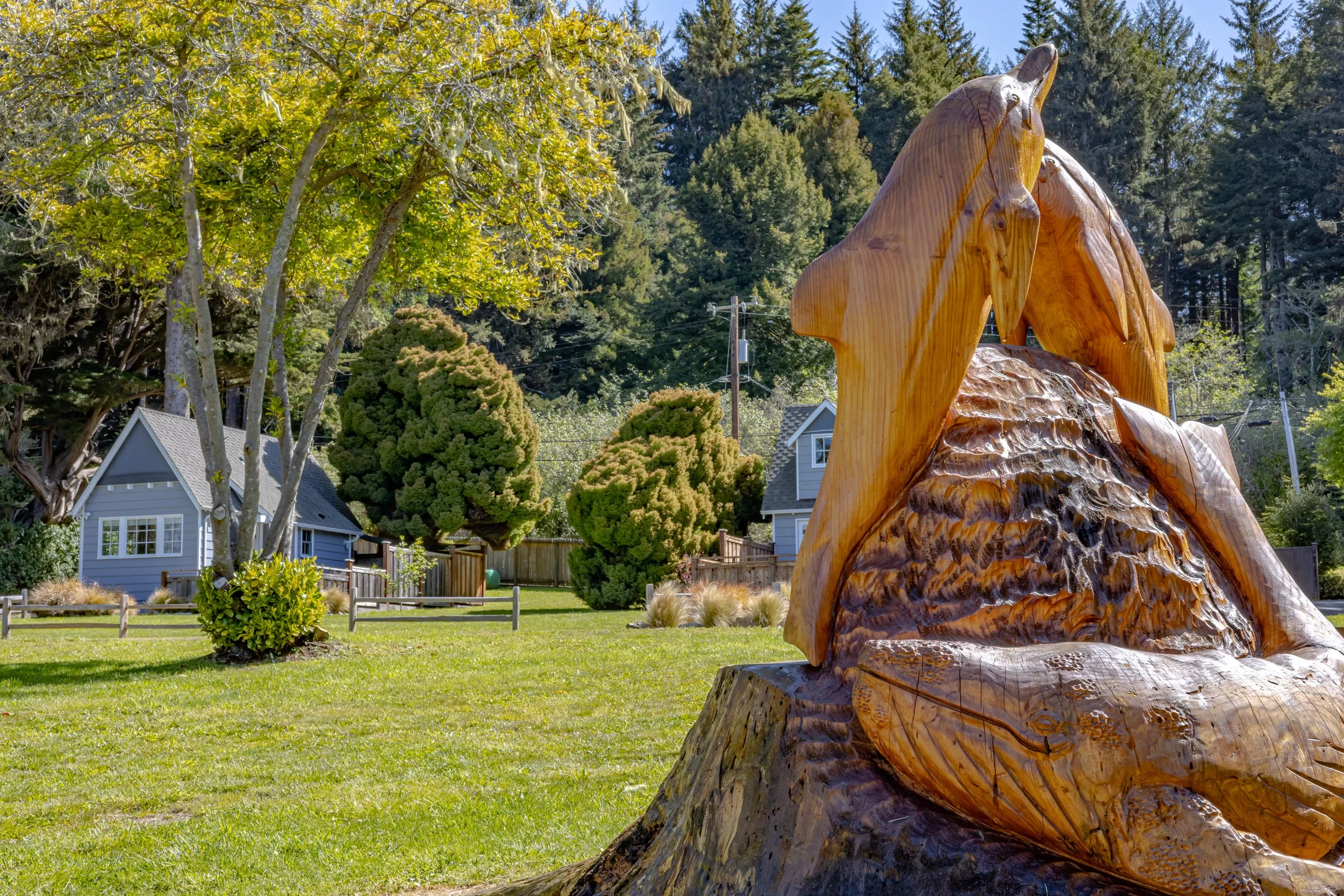 A large wooden sculpture of a bear's head, carved from a tree trunk, located in a lush green backyard with trees, bushes, and a small house in the background.
