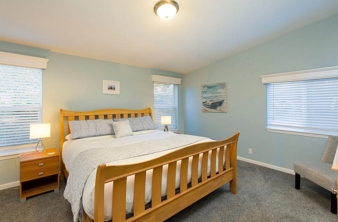 A cozy bedroom with a wooden bed frame, light blue walls, two windows with blinds, a nightstand with a lamp, and a painting of boats on the wall.