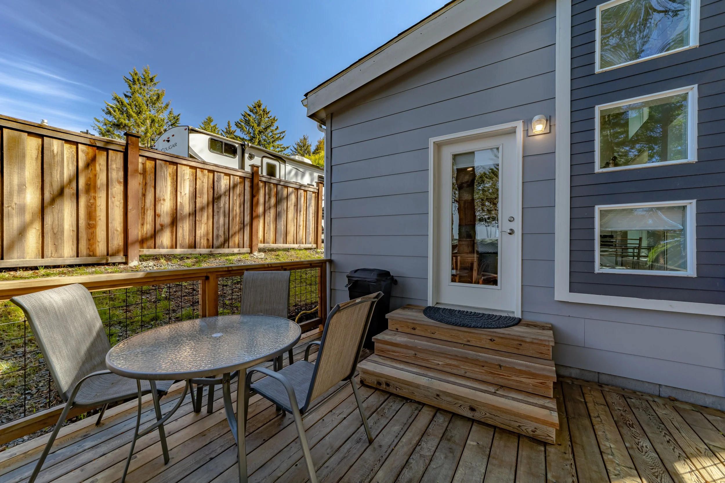Outdoor wooden deck with a round glass table and four chairs, a black trash bin, a sliding glass door, three steps leading to the door, and a wooden fence in a backyard.