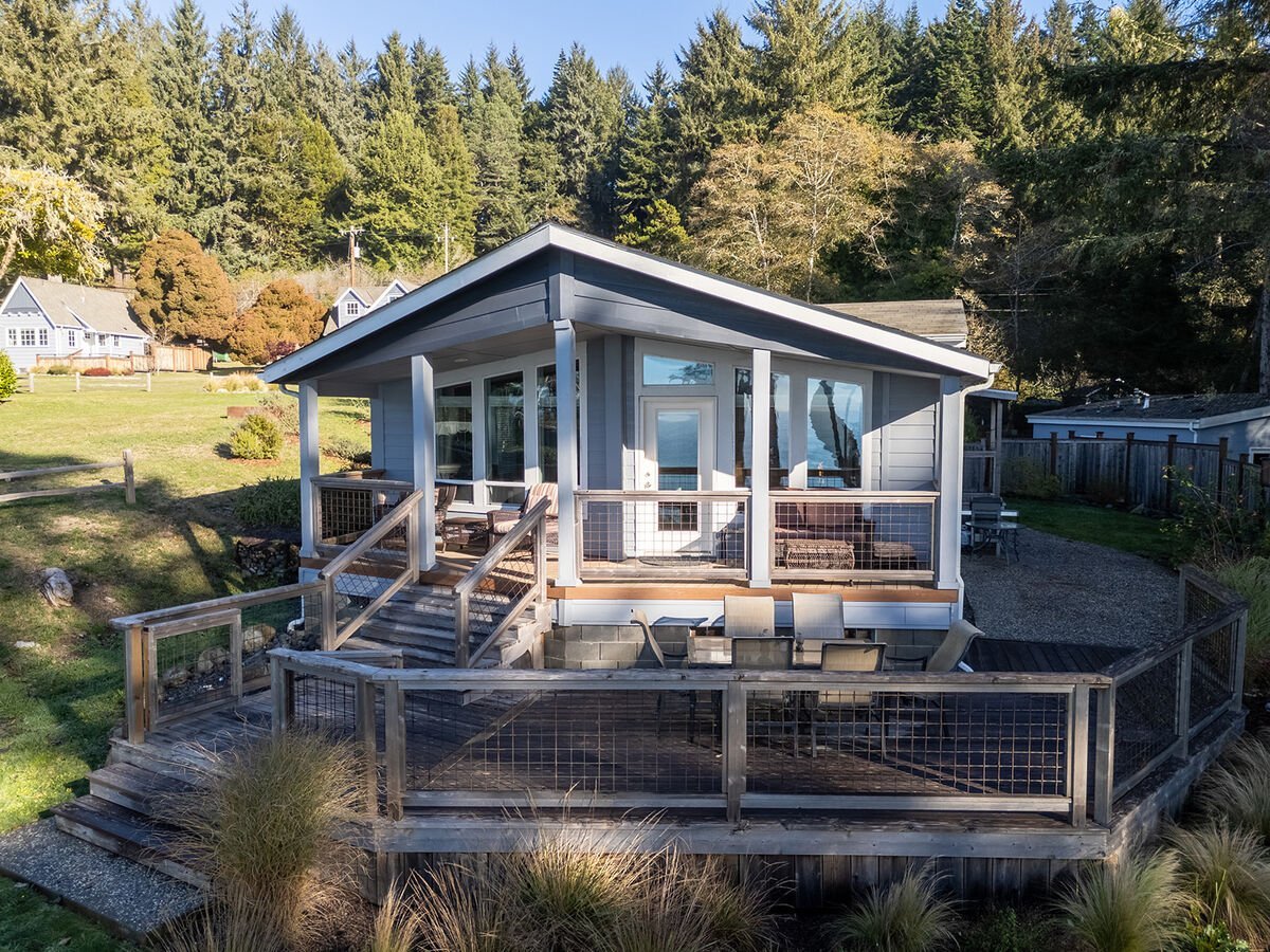 A house with a porch and multiple large windows, surrounded by a fenced deck with outdoor seating, set in a hilly landscape with trees and other houses in the background.