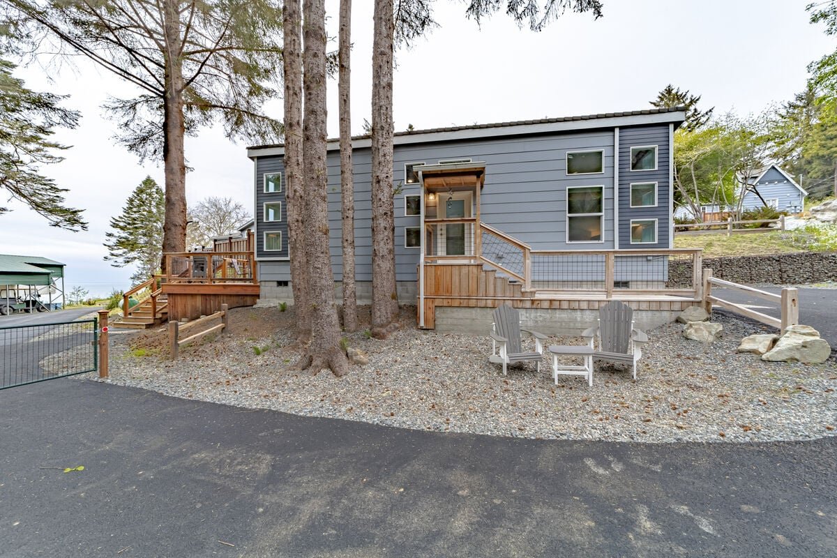 A modern gray house with multiple small windows, surrounded by tall trees, with a wooden deck, two Adirondack chairs, and a small table in the front yard, and an asphalt driveway.