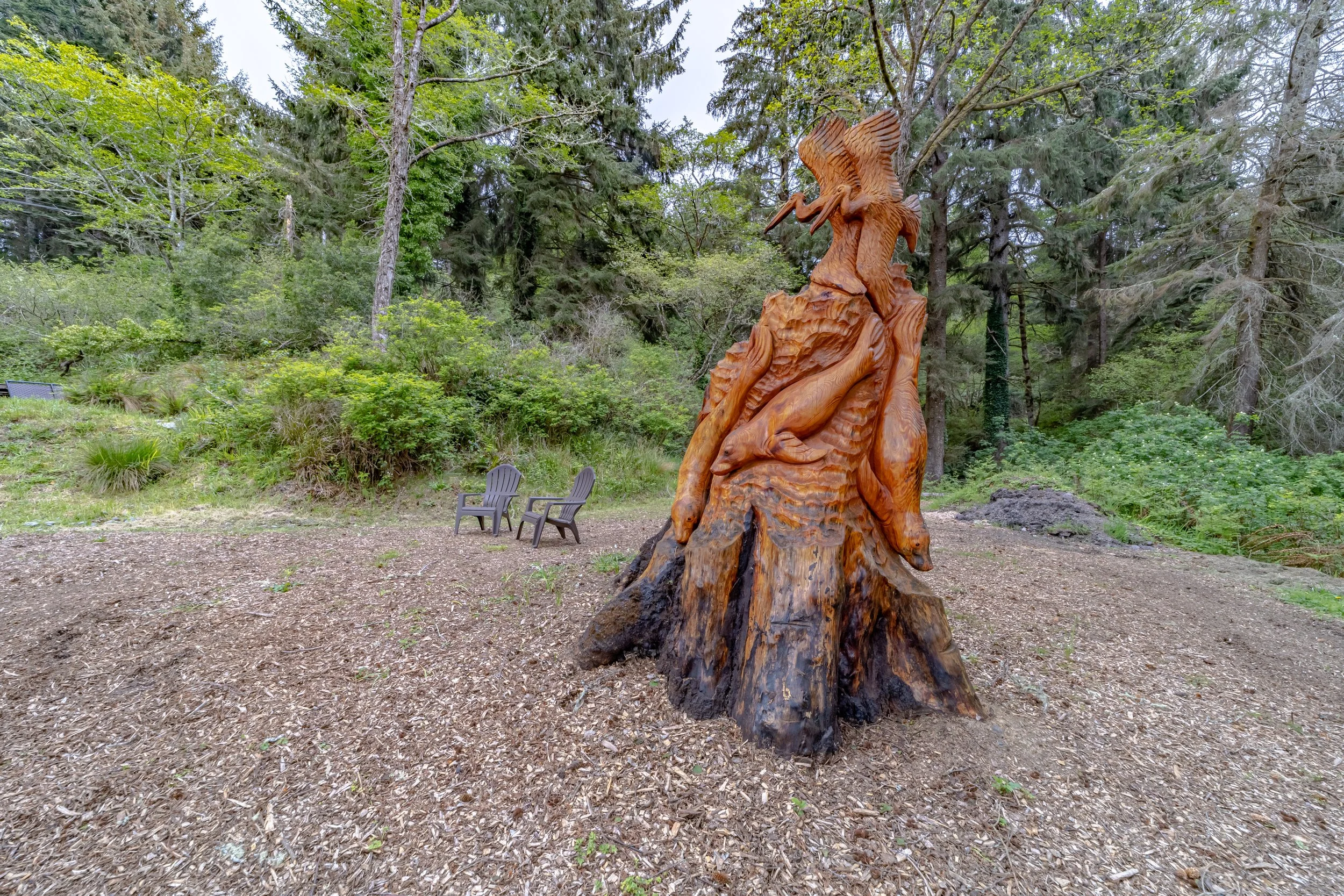 Wooden sculpture of a heron and other animals carved into a large tree trunk, with two outdoor chairs nearby in a forest setting.