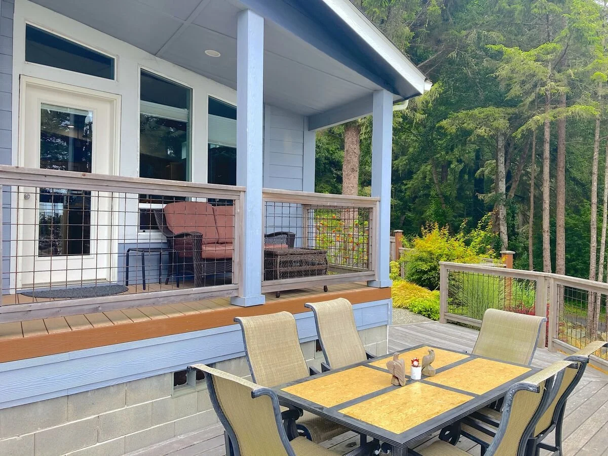 An outdoor deck with a dining table and six chairs, with a house and wooded forest in the background.