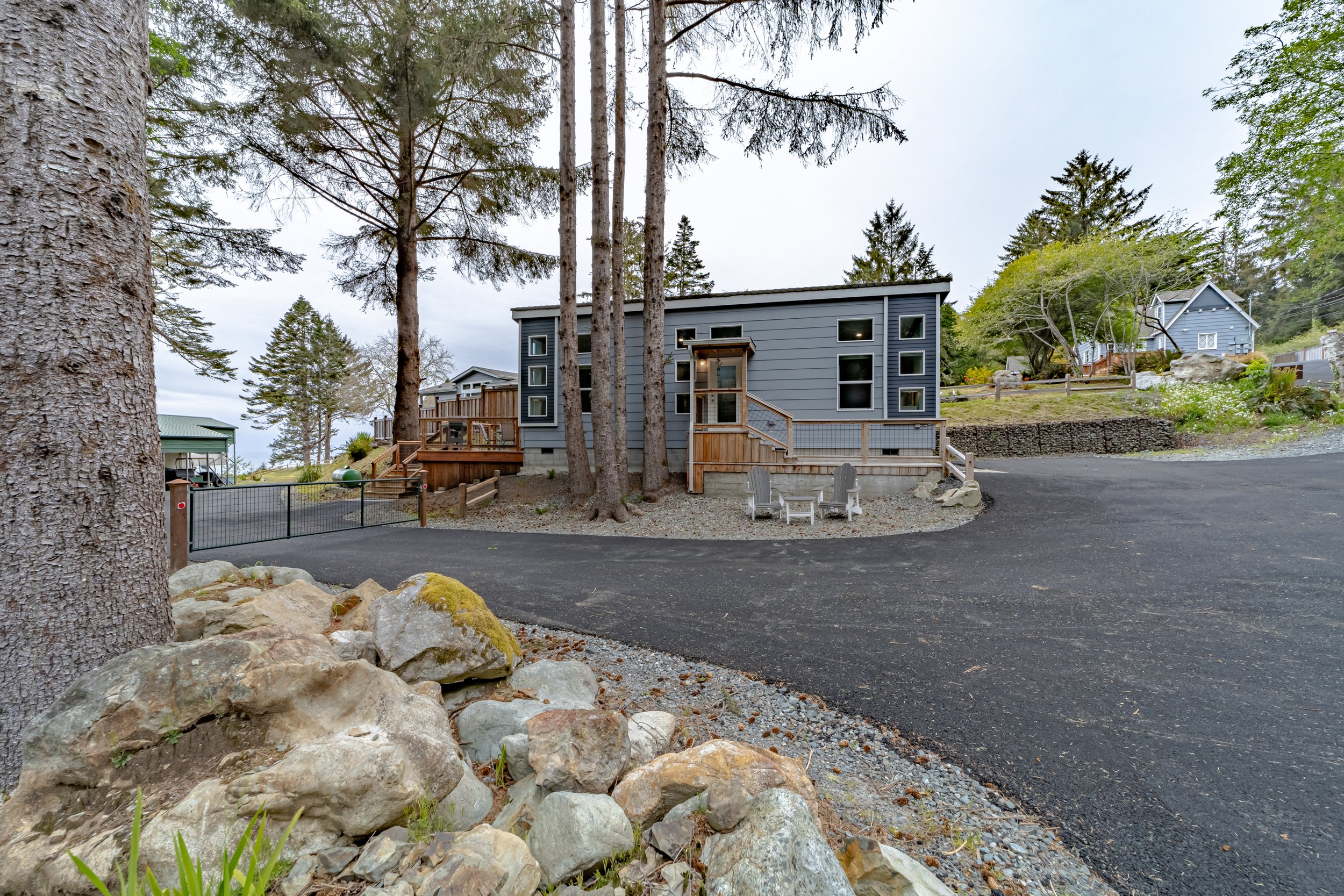 A gray modern house with multiple windows, wooden stairs, and outdoor seating, situated on a gravel driveway surrounded by trees and other houses in a hilly area.