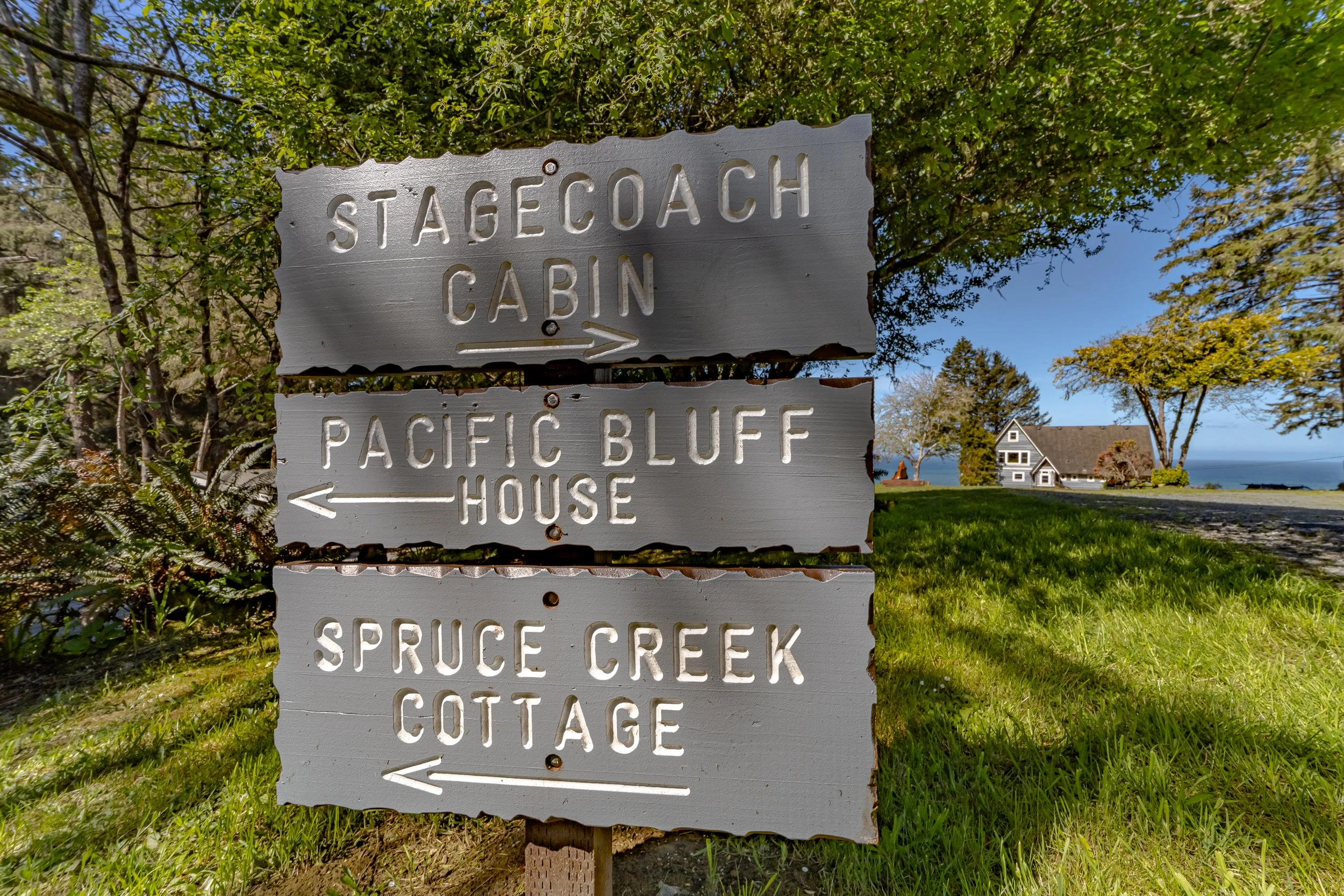 Wooden signpost pointing to three locations: stagecoach cabin, Pacific Bluff House, and Spruce Creek Cottage, with a house and trees in the background.
