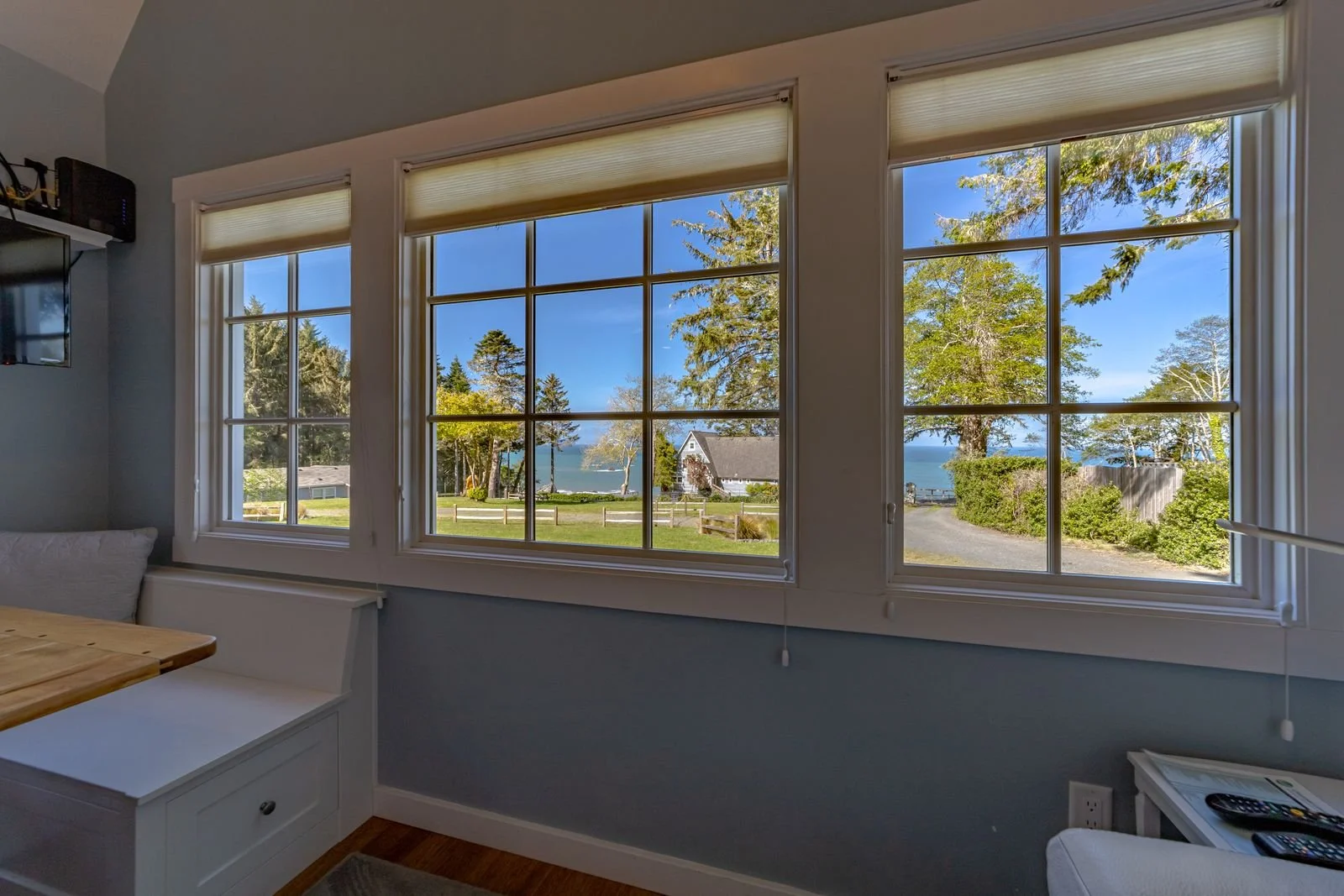 View through three large windows showing trees, a small house, and water outside, in a cozy room with blue walls, a wooden table, and remote controls on a white side table.