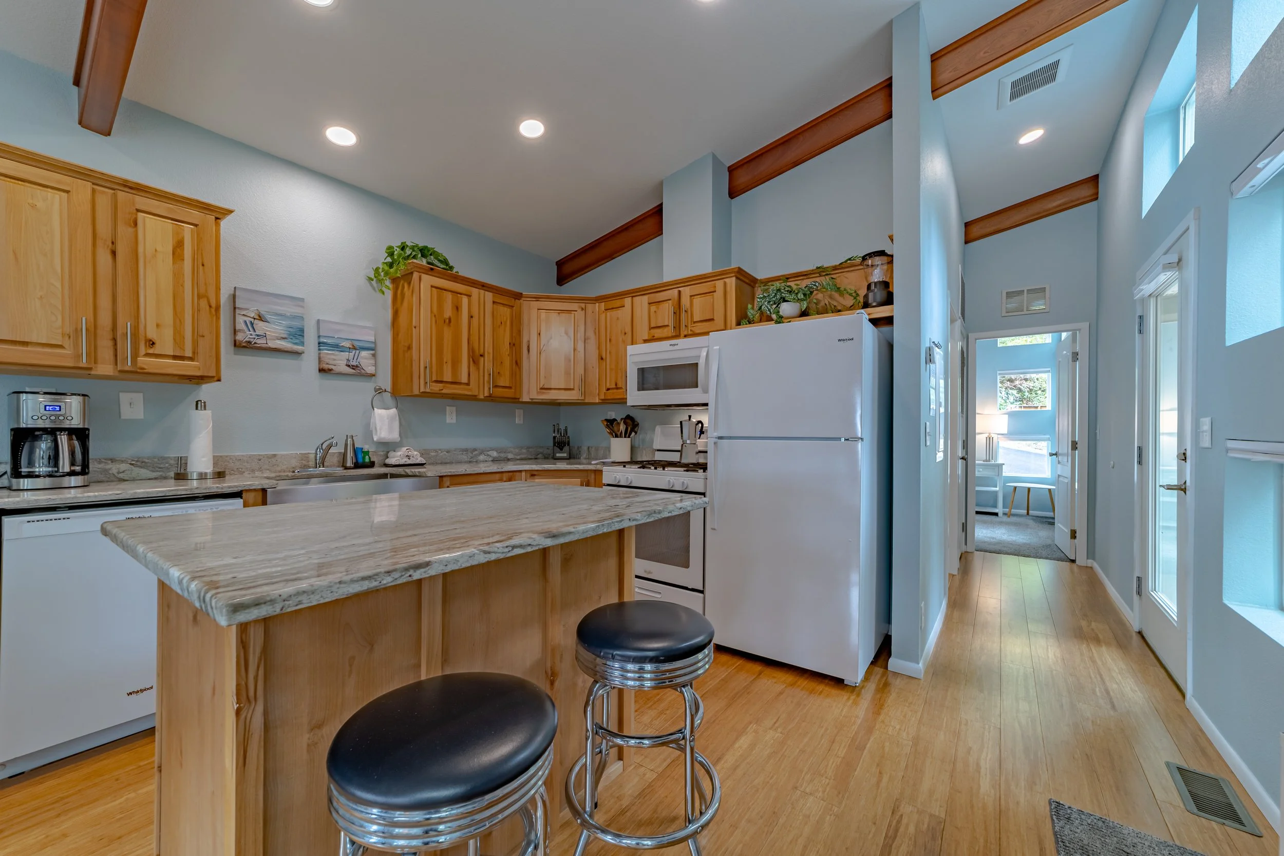A cozy kitchen with wooden cabinets, a marble countertop island, white appliances, and hardwood floors, with a view into a bright adjacent room.