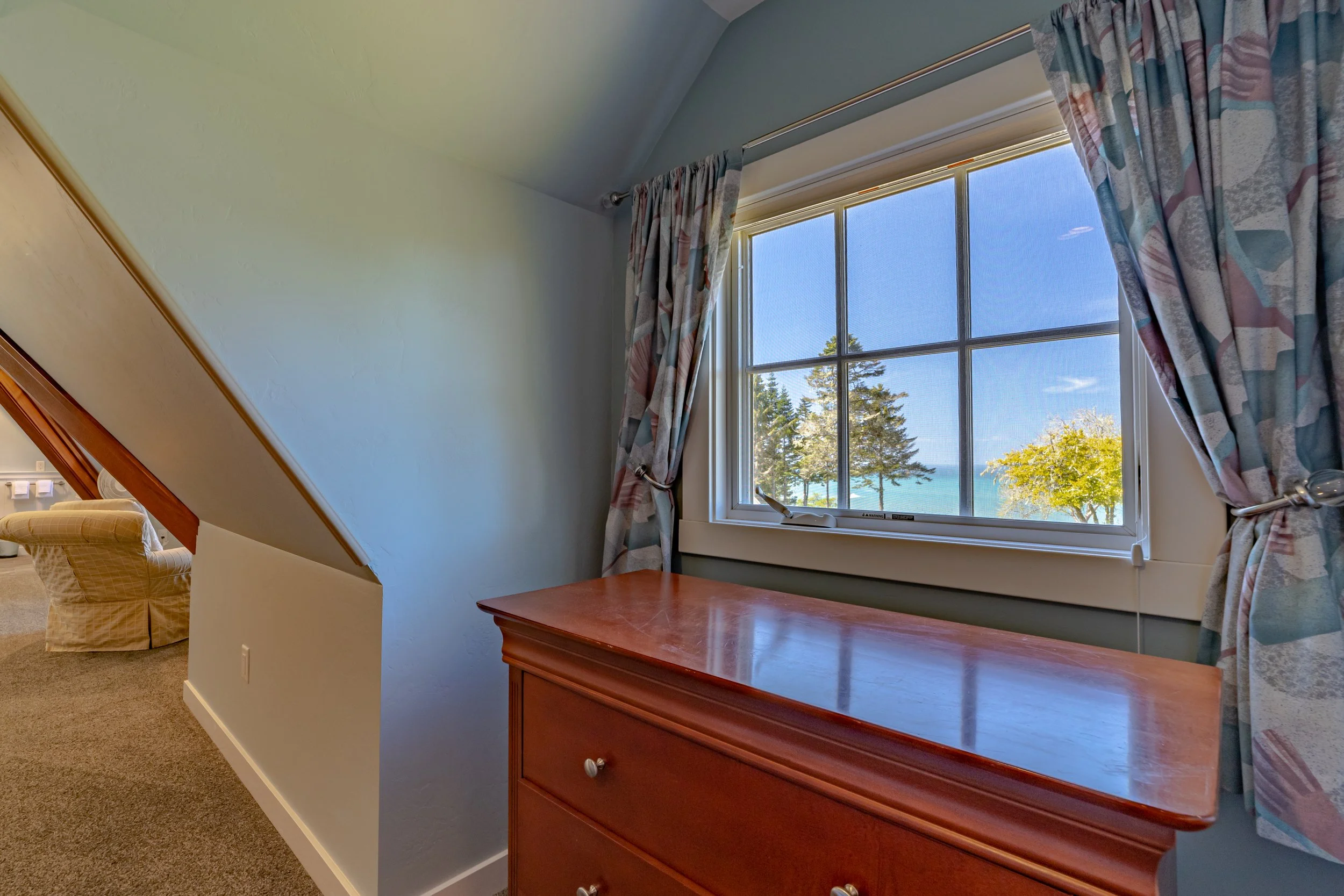 Interior of a room with a window showing trees and water outside, surrounded by blue sky. There is a wooden dresser below the window and curtains on either side. Part of a beige armchair is visible in the adjacent room.