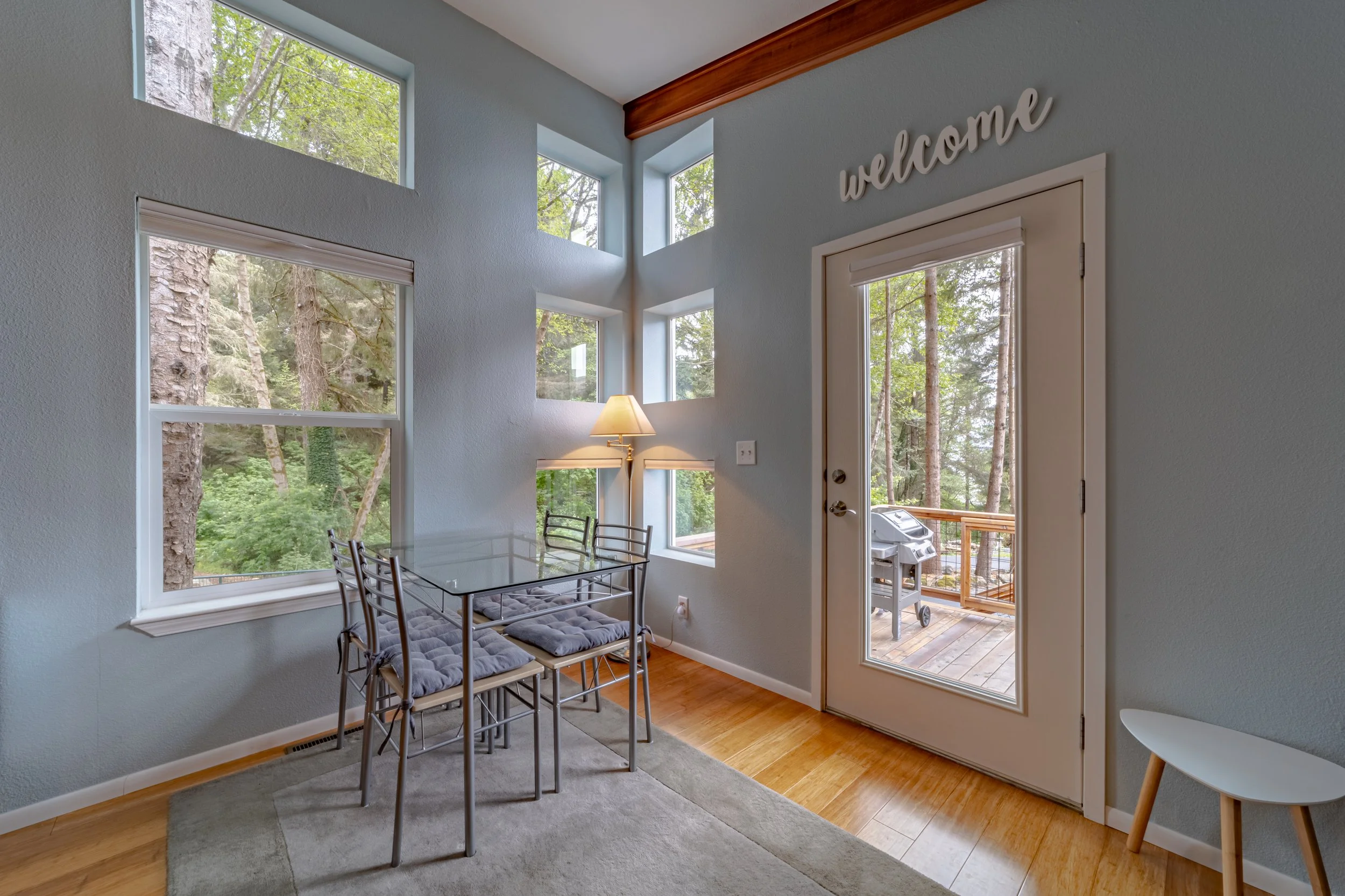 Interior of a dining area with a glass table and four chairs, large windows showing trees outside, a wall lamp, a door leading to a balcony with a grill, and a 'welcome' sign on the wall.