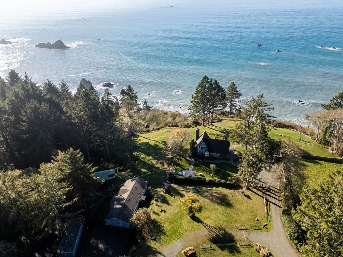 Aerial view of a coastal property with a house, surrounded by trees, near the ocean with rocks and waves.