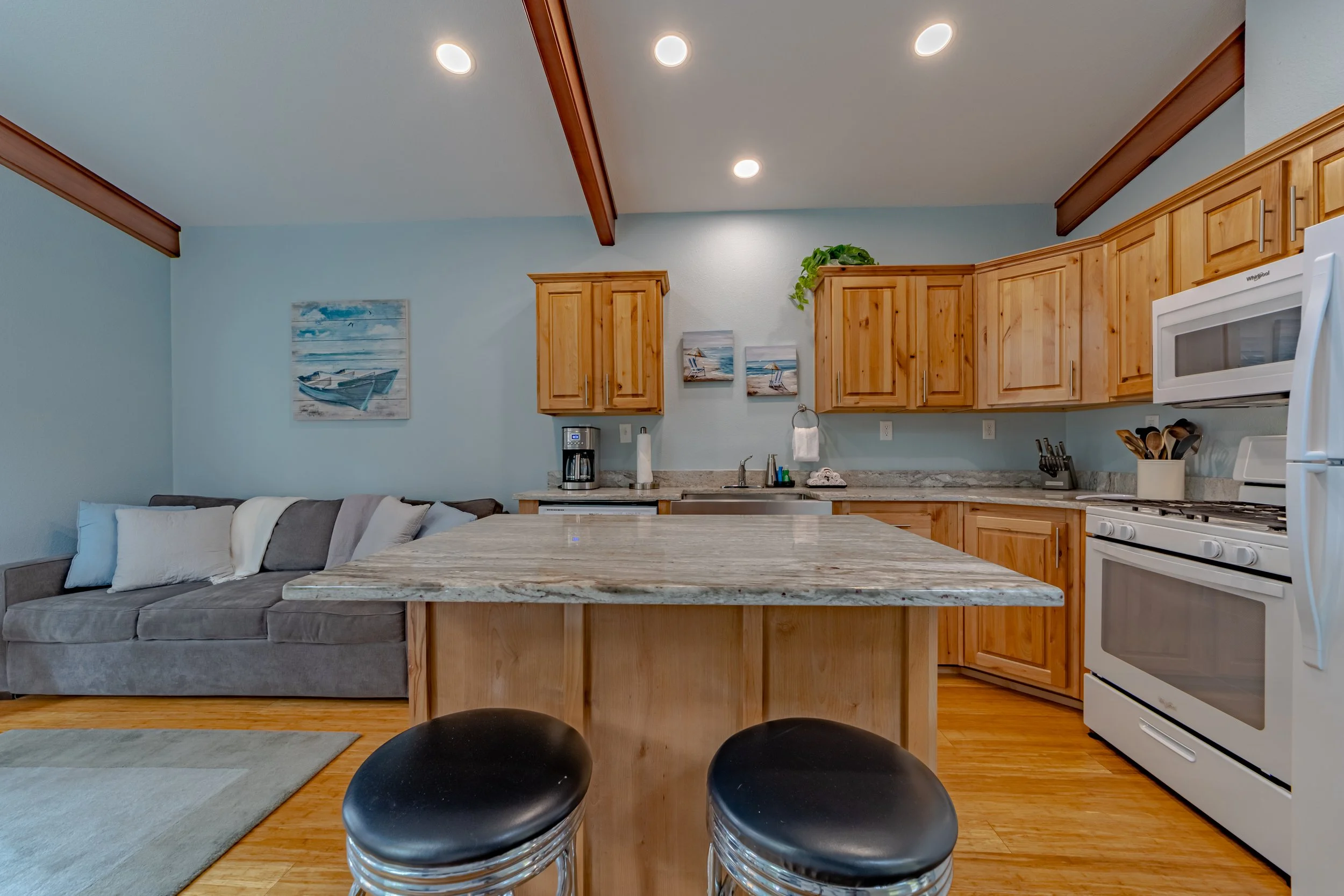 Kitchen with wooden cabinets, marble countertop island, white appliances including a stove and microwave, coffee maker, and a living area with a gray couch and pillows.