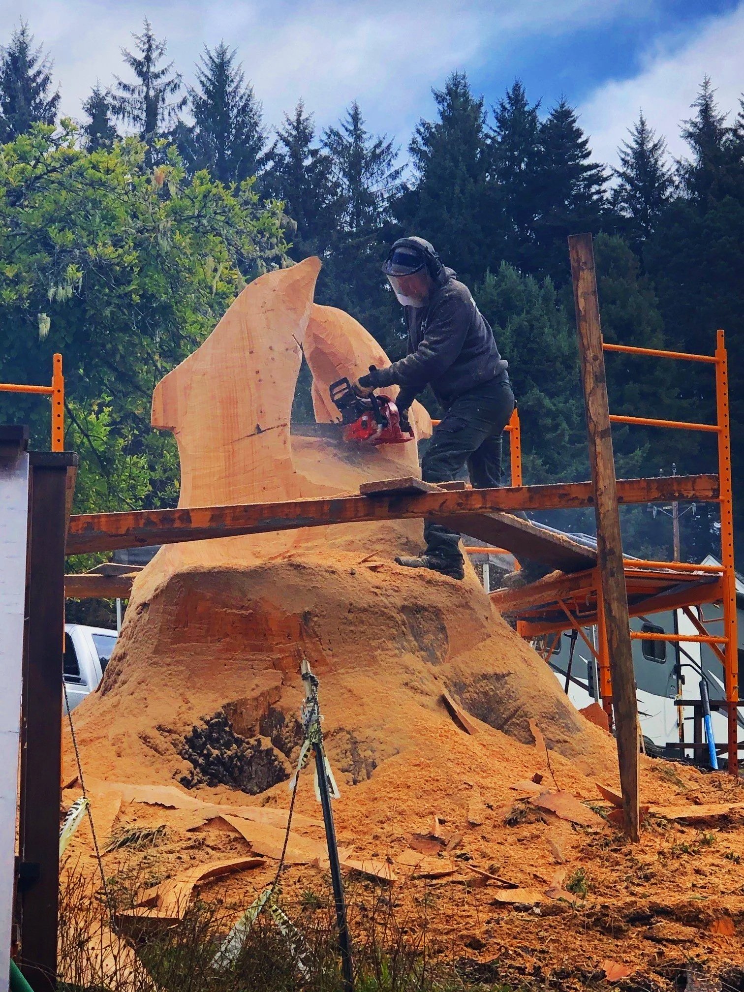 A person wearing safety gear, including a face shield and headphones, is using a chainsaw to carve or shape a large wooden sculpture in an outdoor workshop surrounded by tall trees and blue skies.