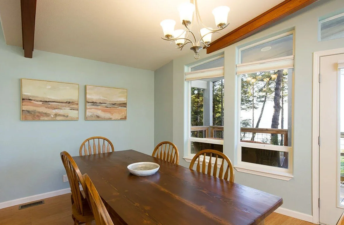 Wooden dining table with four chairs in a bright room with large windows overlooking trees, a chandelier on the ceiling, and wall paintings.