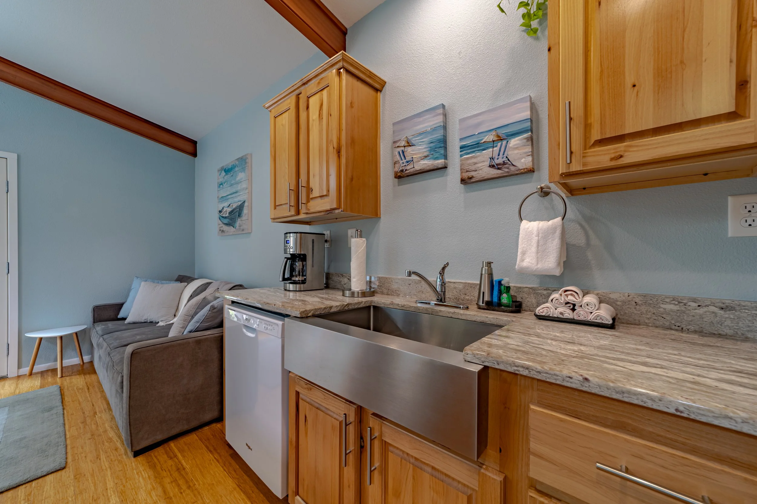 Kitchen with wooden cabinets, granite countertops, stainless steel farmhouse sink, coffee maker, paper towel holder, and rolled-up towels. Part of a living room with a gray sofa and wall art featuring beach scenes.