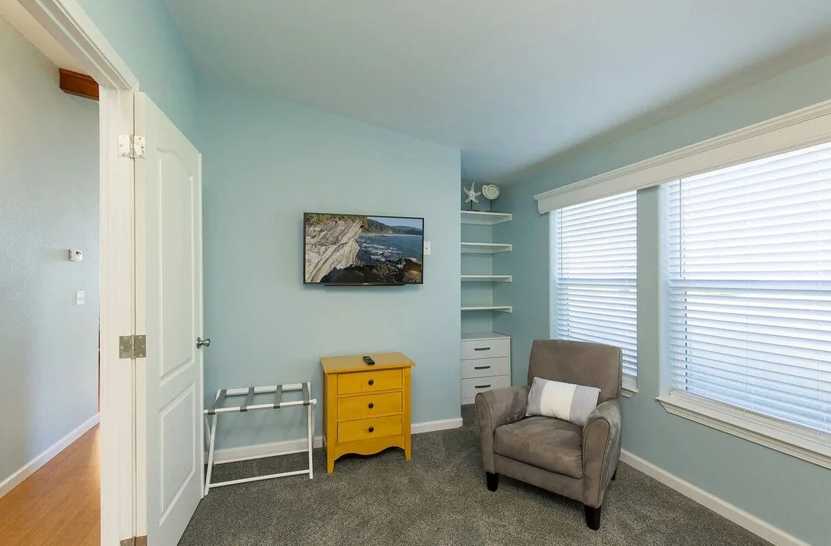 Living room with blue walls, a gray armchair with a white pillow, a yellow side table, white shelving, and three large windows with blinds.