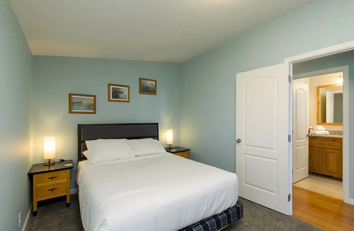 A bedroom with a king-sized bed, two nightstands with lamps, light blue walls, framed pictures above the bed, and an open door leading to a bathroom with a wooden vanity and mirror.