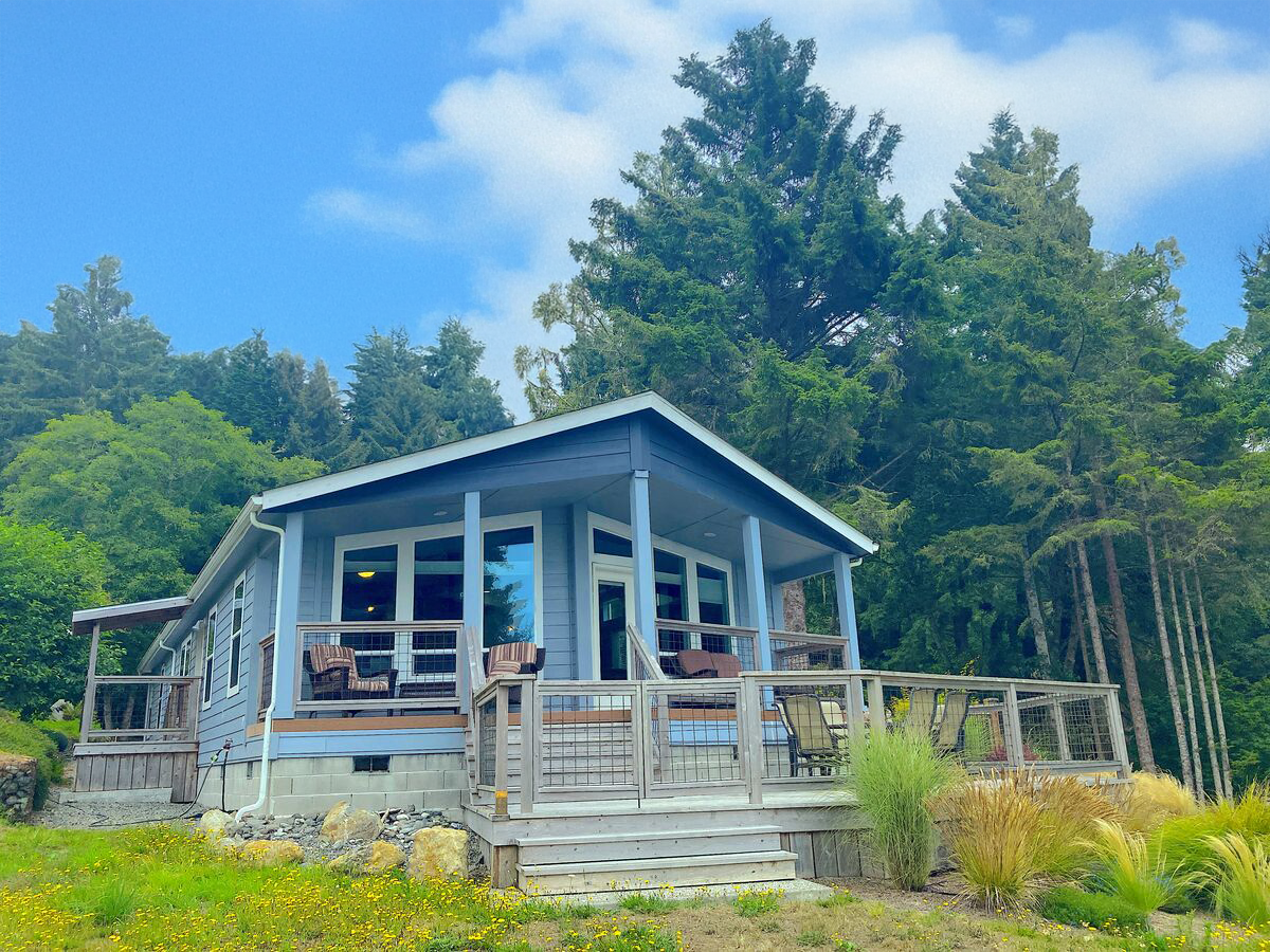 A light blue house with a porch, railing, and outdoor furniture, set against a backdrop of tall green trees and a partly cloudy sky.