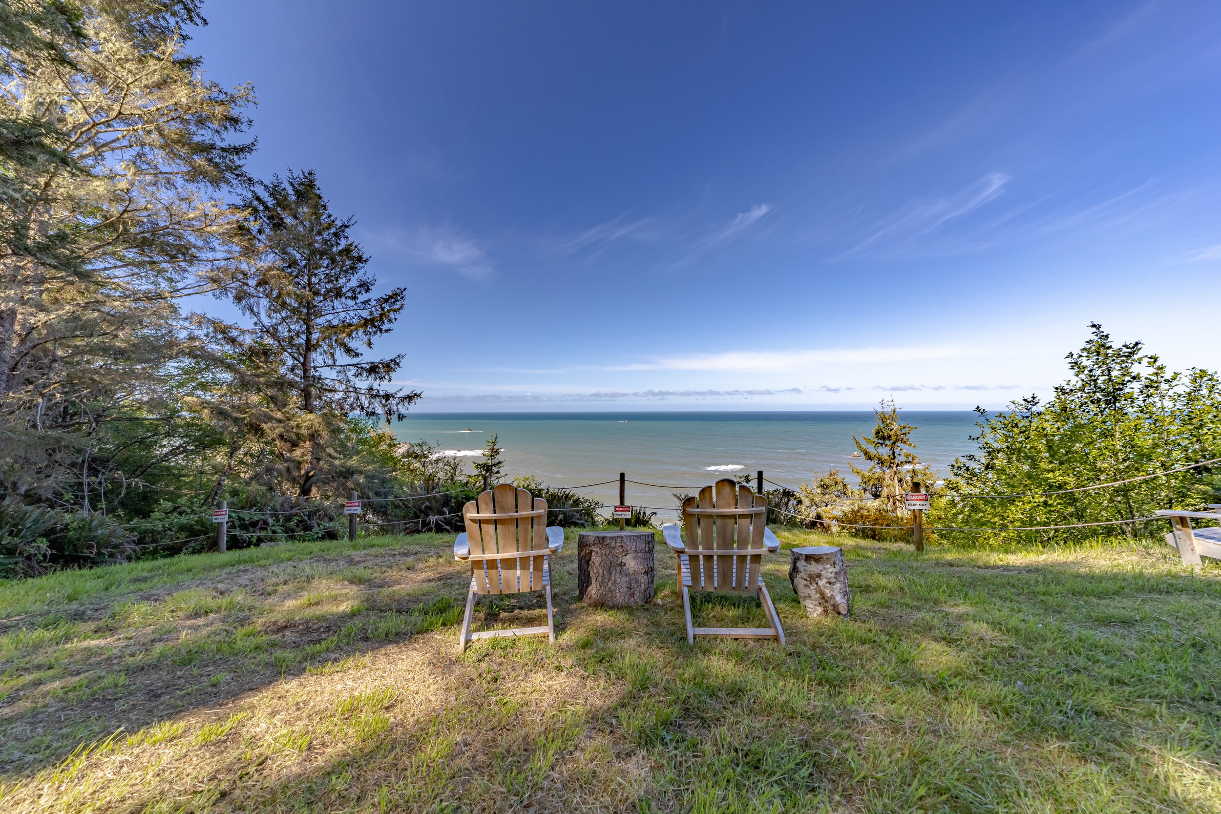 Two wooden Adirondack chairs and two tree stumps facing the ocean on a grassy hill, with trees and a blue sky in the background.