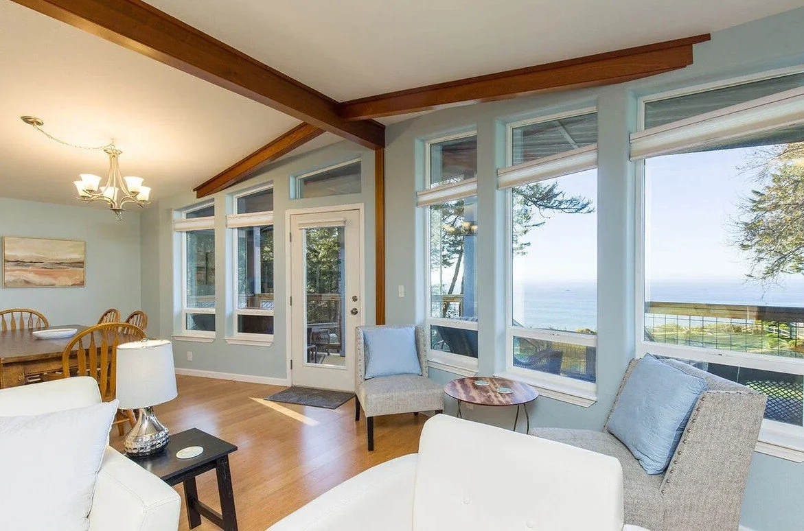 Living room with large windows overlooking a scenic view of trees and water, furnished with chairs, a table, and a chandelier.
