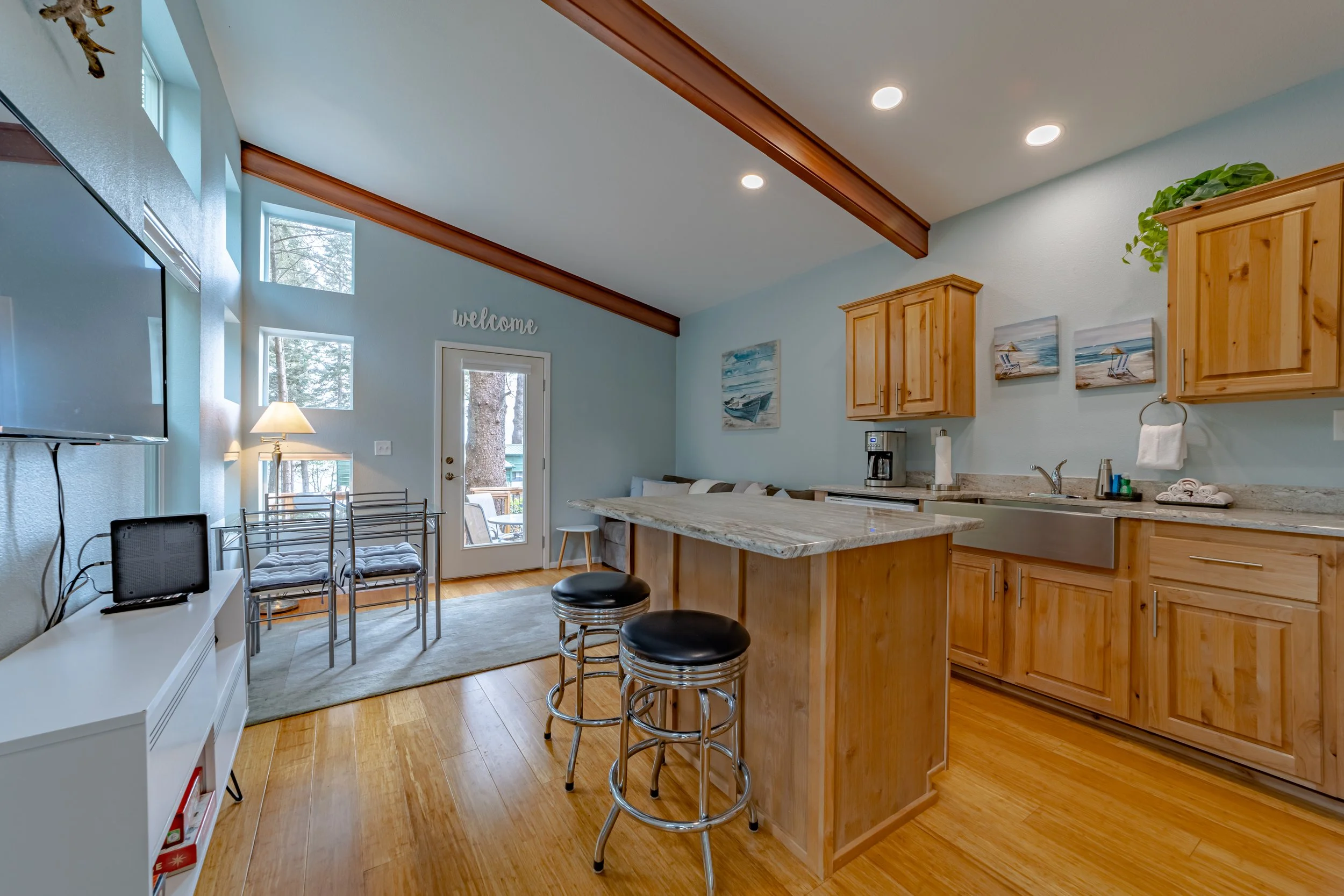 Open-concept kitchen and living area with light blue walls, wooden cabinets, and hardwood floors. There is a kitchen island with two black stools, a sink, a coffee maker, and three paintings of beach scenes on the wall. A sliding glass door leads to 