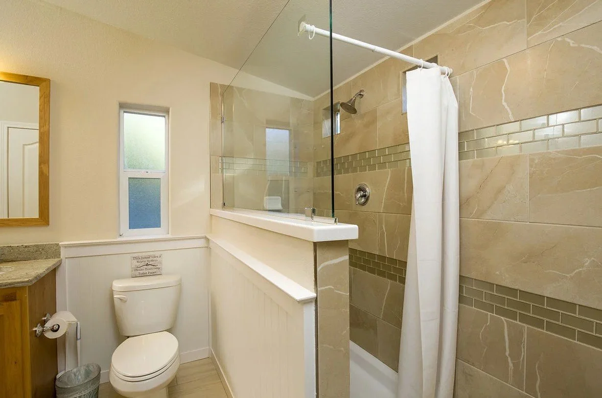 A bathroom with a toilet, a small window, a mirror, and a walk-in shower with beige tiles and a white curtain.