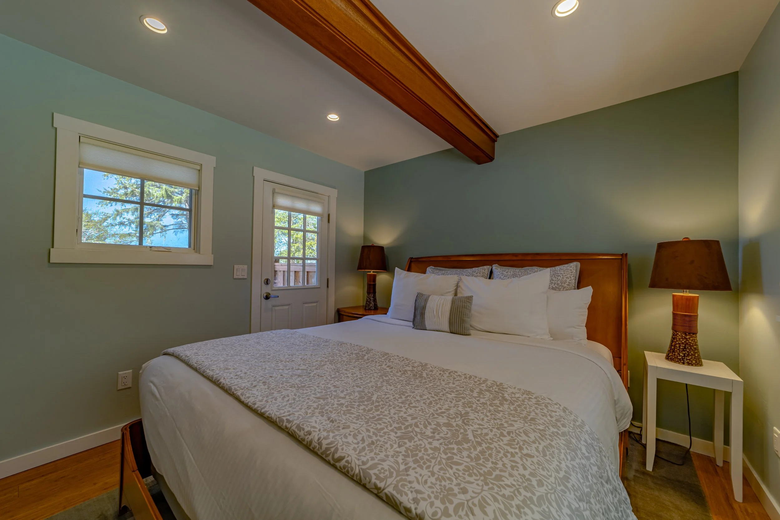 Cozy bedroom with a large bed, multiple pillows, two bedside lamps, a small window, and a door with a window, with light blue walls and hardwood floors.