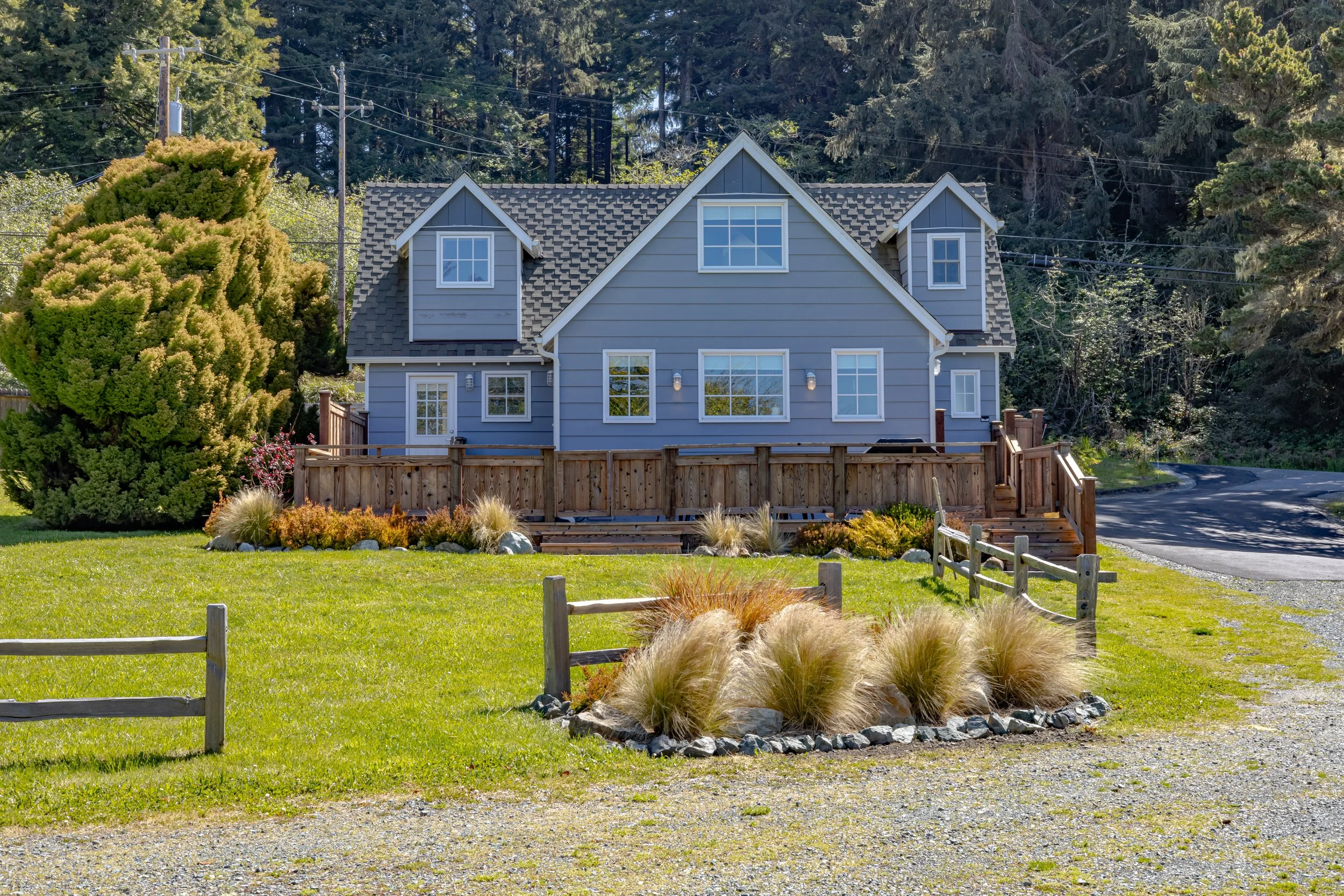 A two-story blue house with white trim, multiple windows, and a gabled roof, surrounded by green lawn and landscaping, with large trees in the background.