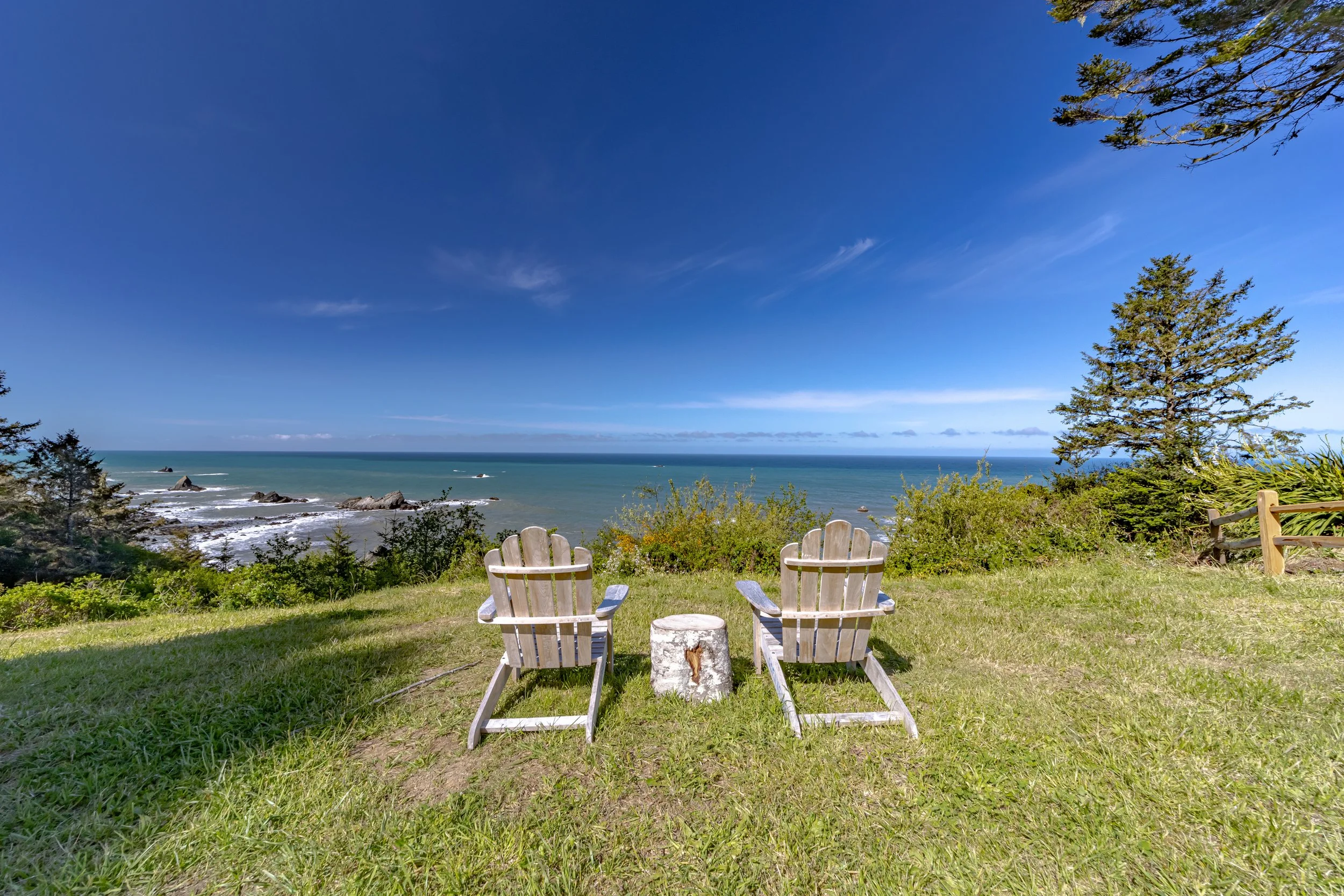 Two Adirondack chairs facing the ocean with a small log in between, overlooking a grassy slope, trees, and a blue sky with some clouds.