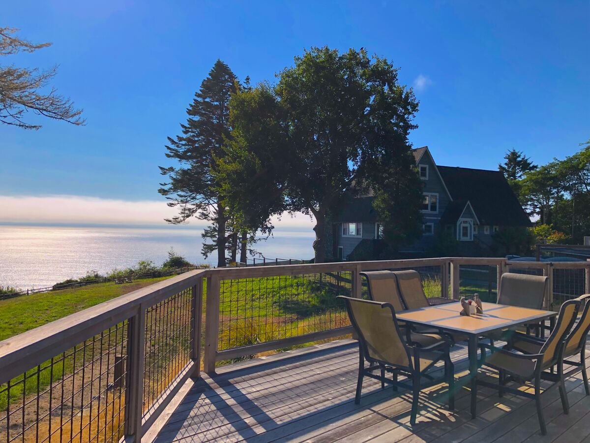 A wooden deck with a table and six chairs overlooking a body of water, with a house and trees in the background, under a blue sky.