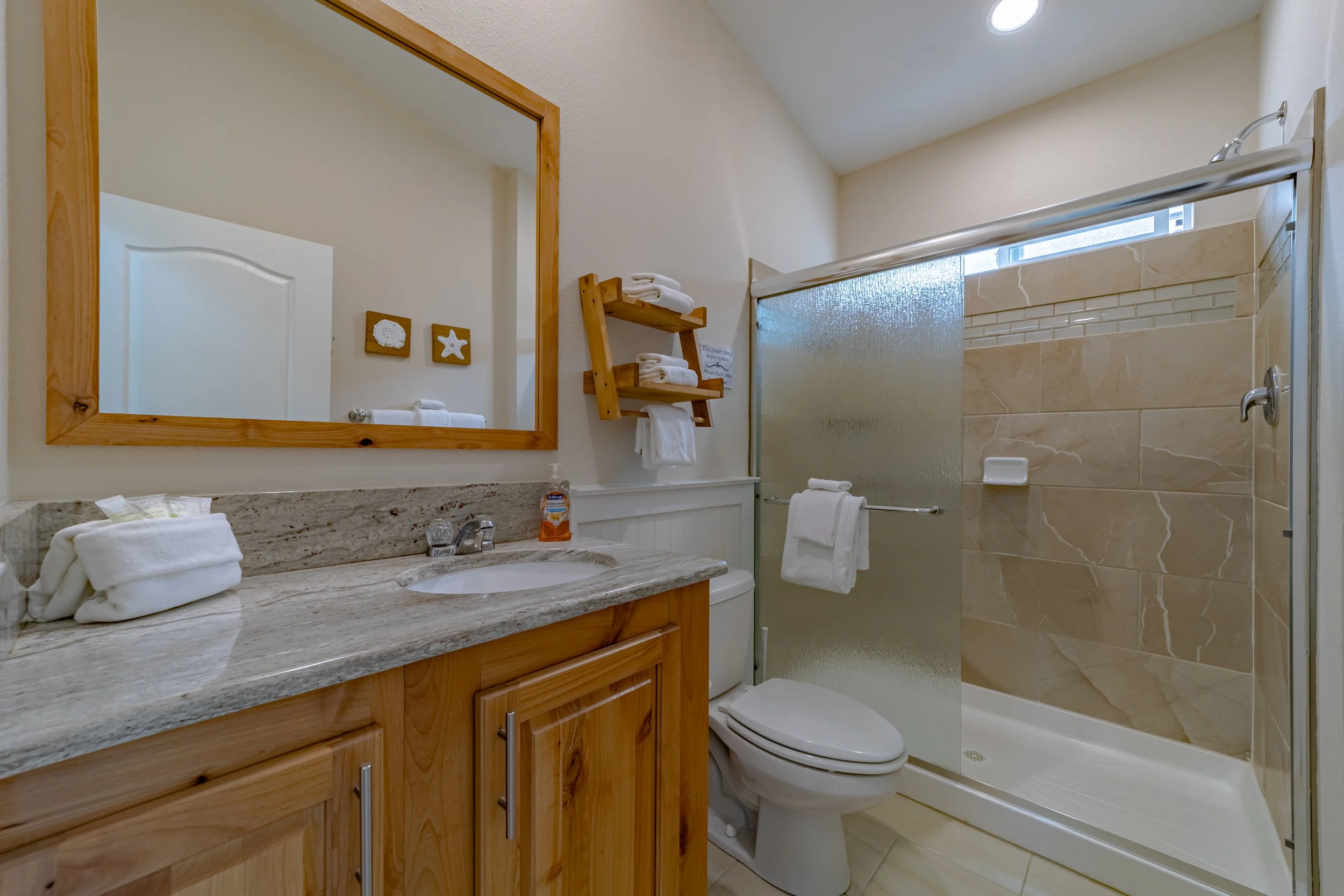 Bathroom with wooden vanity, granite countertop, mirror, towel rack, glass shower enclosure with tan tiles, and decorative wall art.