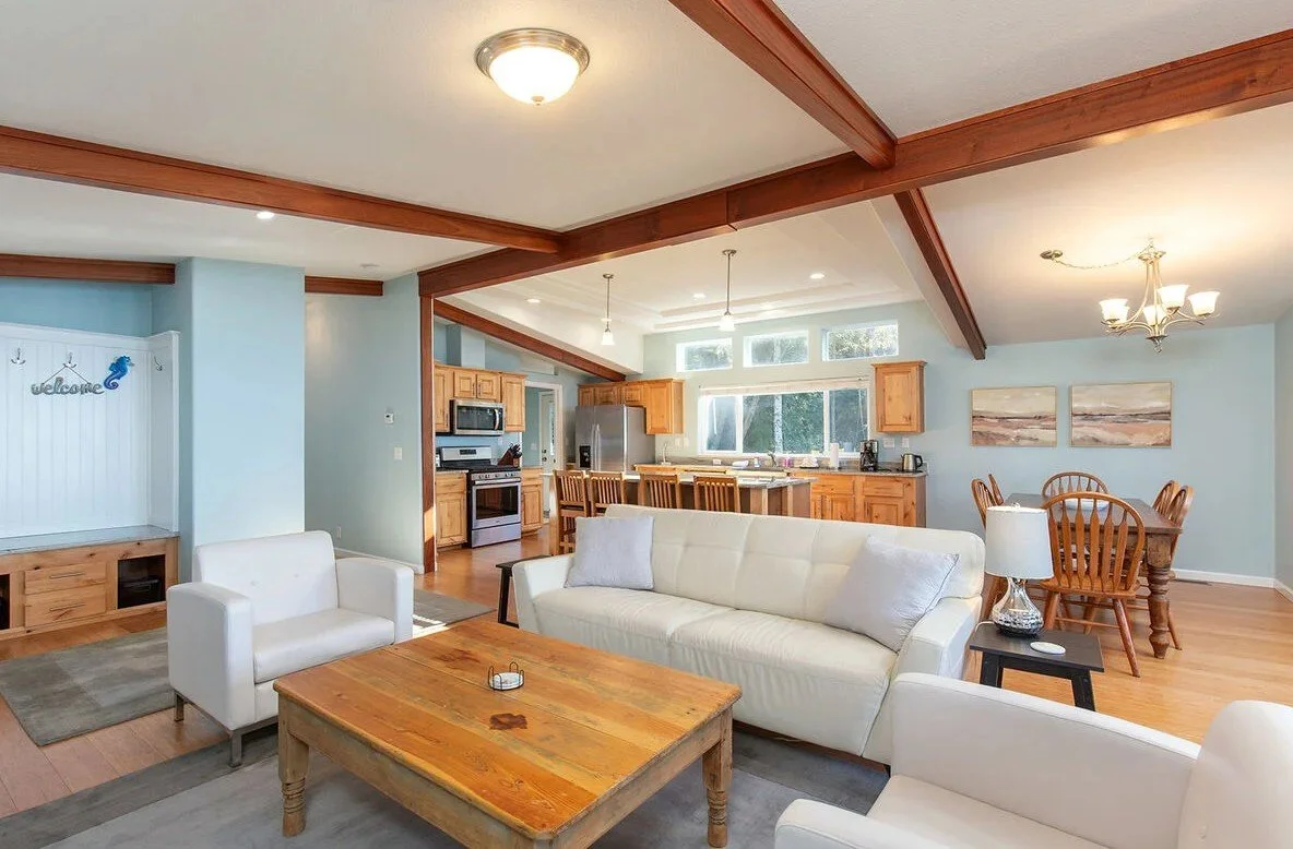 Living room with a white sofa, armchair, wooden coffee table, and a lamp, open to a kitchen and dining area with wooden furniture, large windows, and ceiling lights.