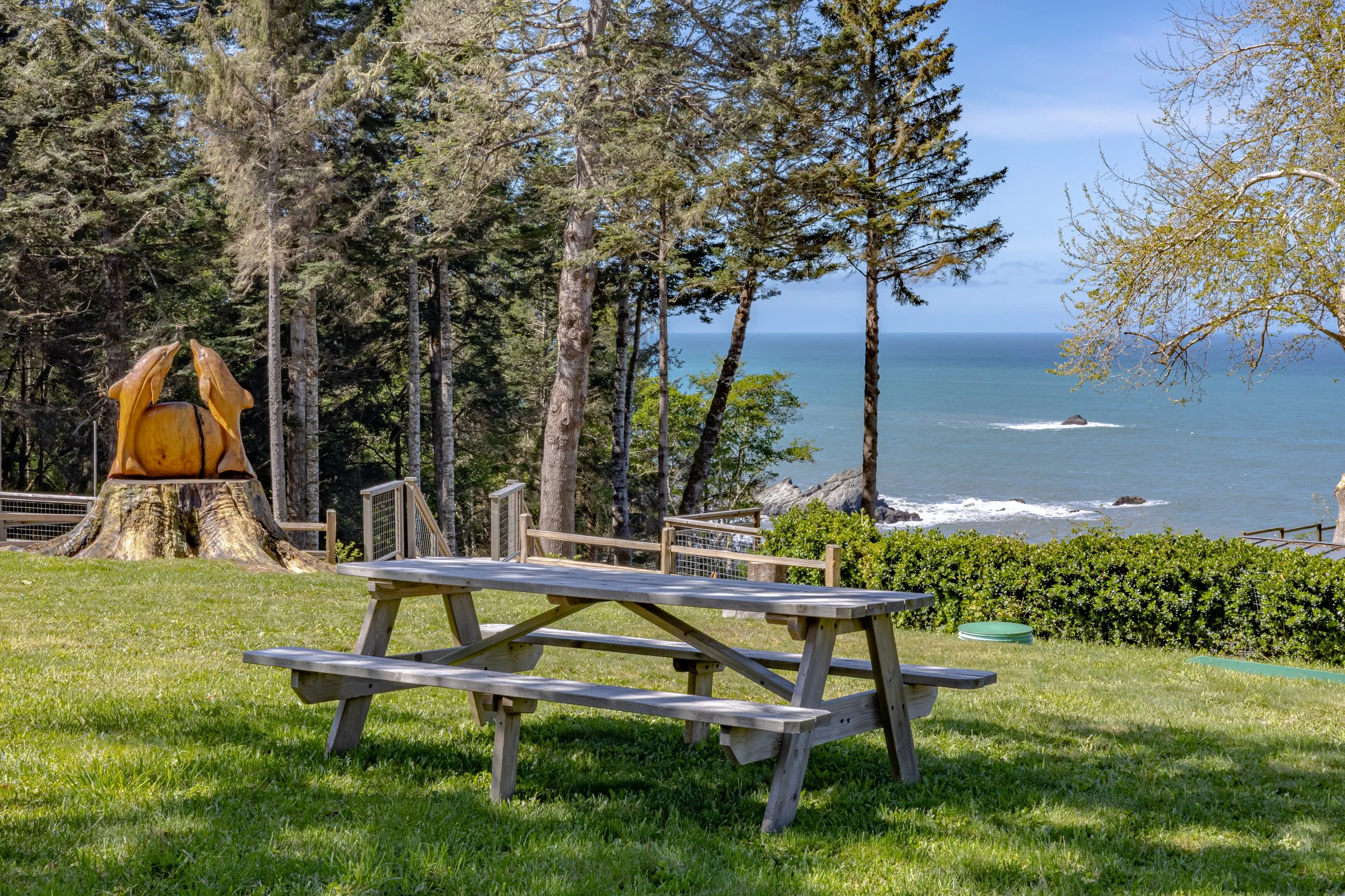 Picnic table on a grassy area near a forest and ocean view, with trees, a tree stump sculpture, bushes, and rocks in the background.